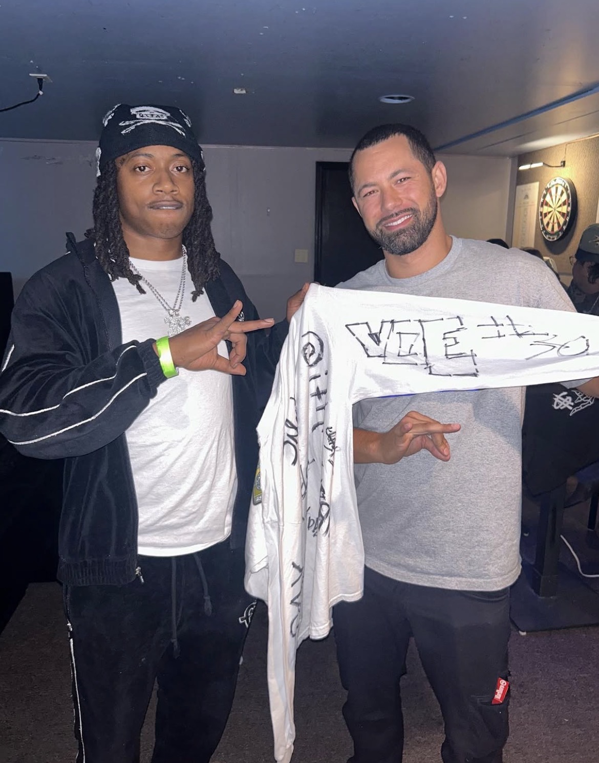 Two men standing indoors, one holding a large white cloth with writing, both smiling at the camera.