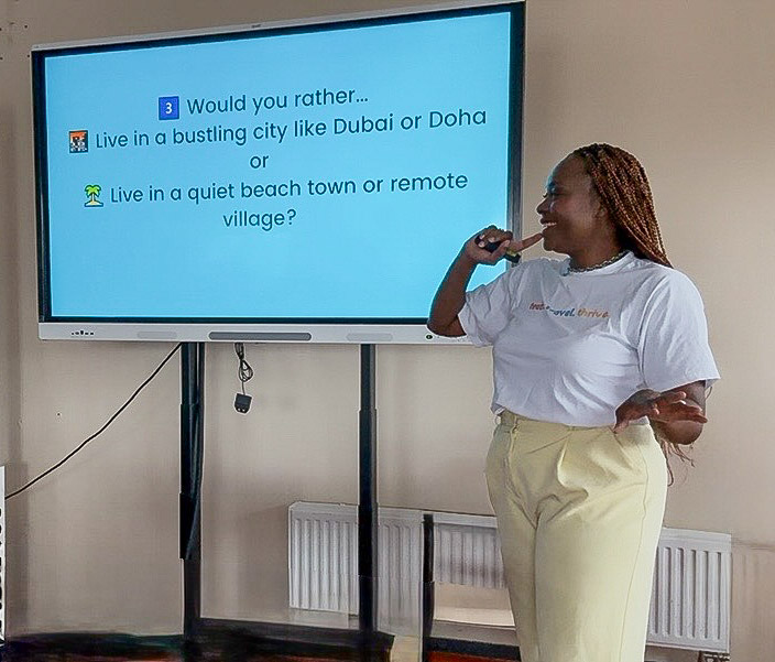 Woman standing next to a screen, holding a microphone, wearing a white shirt and yellow pants, in a room with a radiator.
