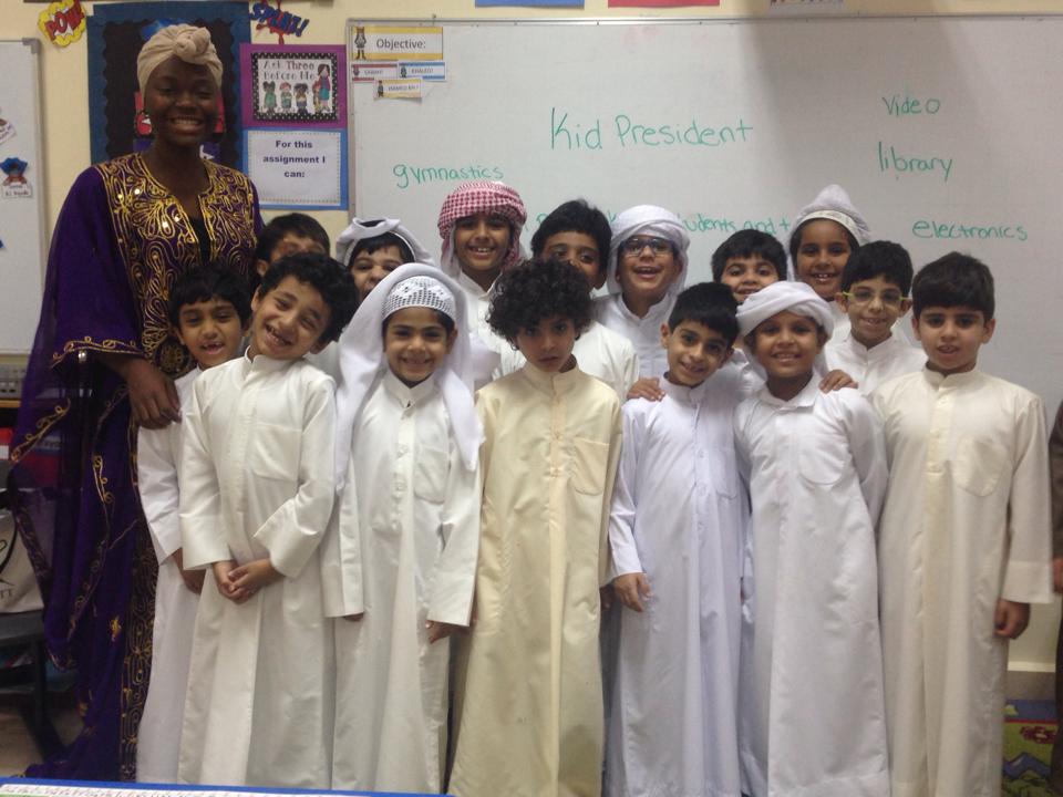 Group of children in white traditional clothing with a woman in colorful attire, standing in classroom in front of a whiteboard.