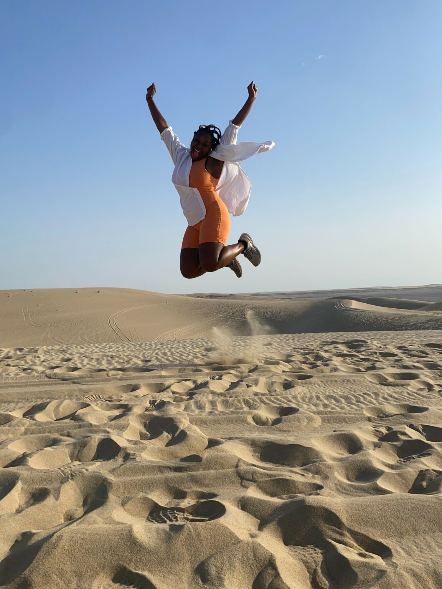 Person jumping in the air on sandy desert with clear blue sky, arms raised, wearing orange dress and white shirt.