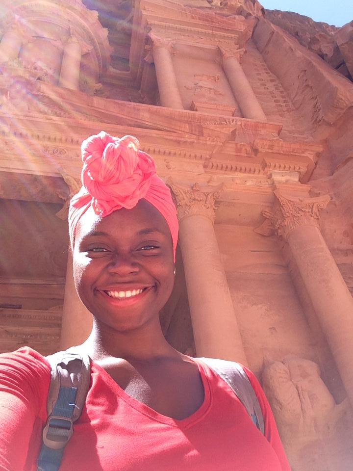 Smiling woman wearing a pink headwrap and red shirt in front of ancient stone structure.
