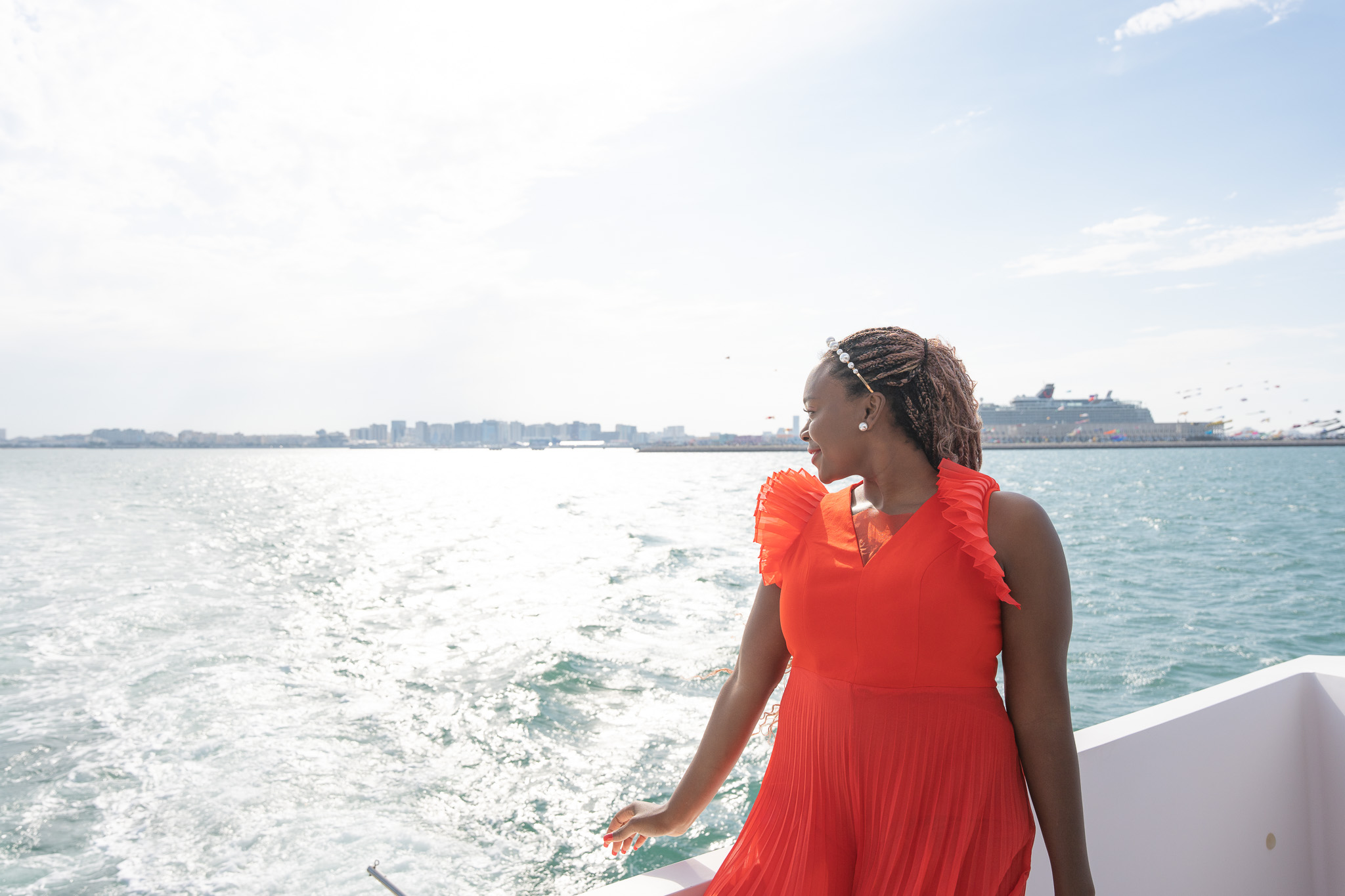 Woman in red dress standing near water with city skyline in background, sunny day.