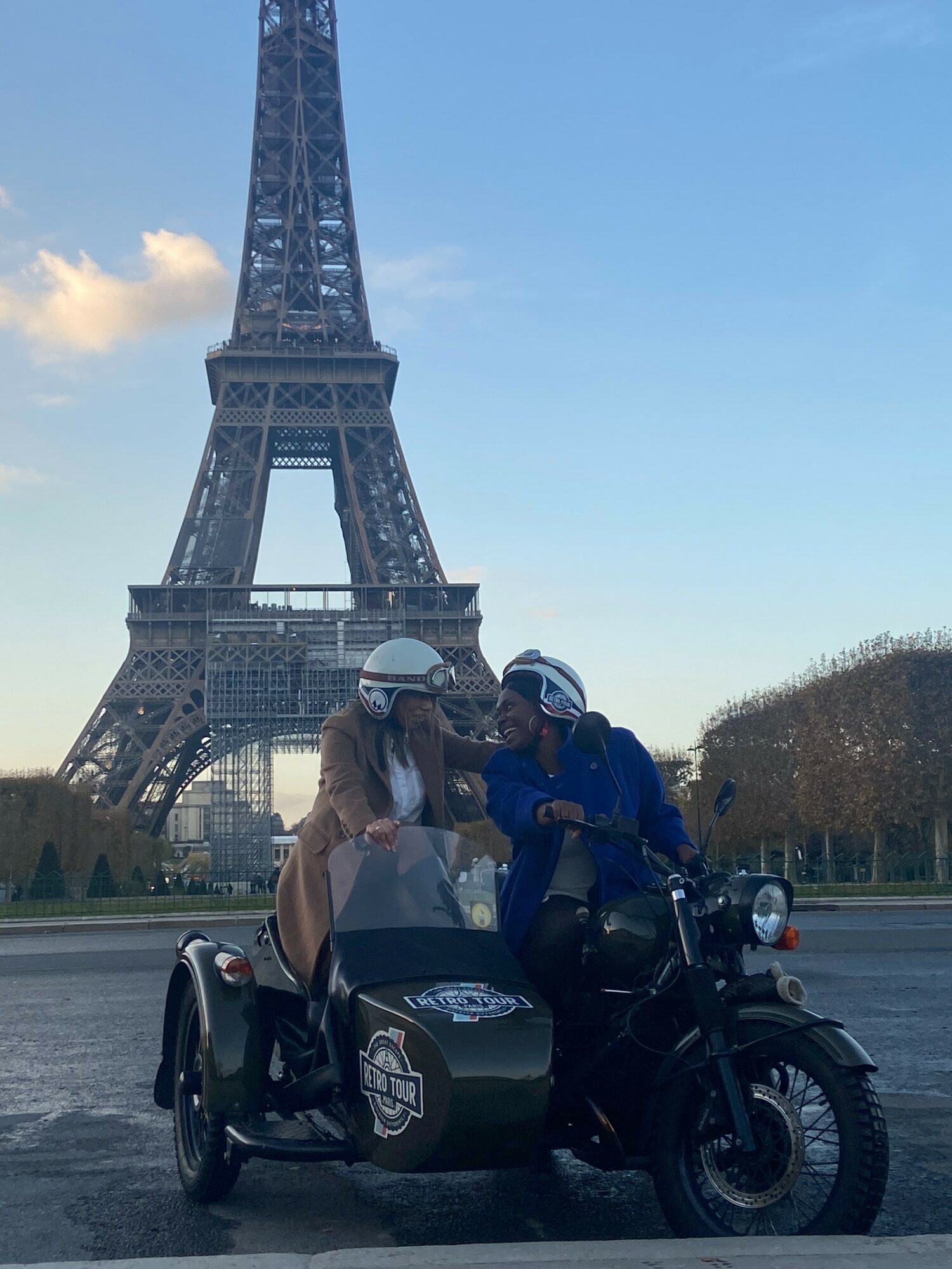 Two people on a motorcycle with a sidecar in front of the Eiffel Tower, talking, during daytime.