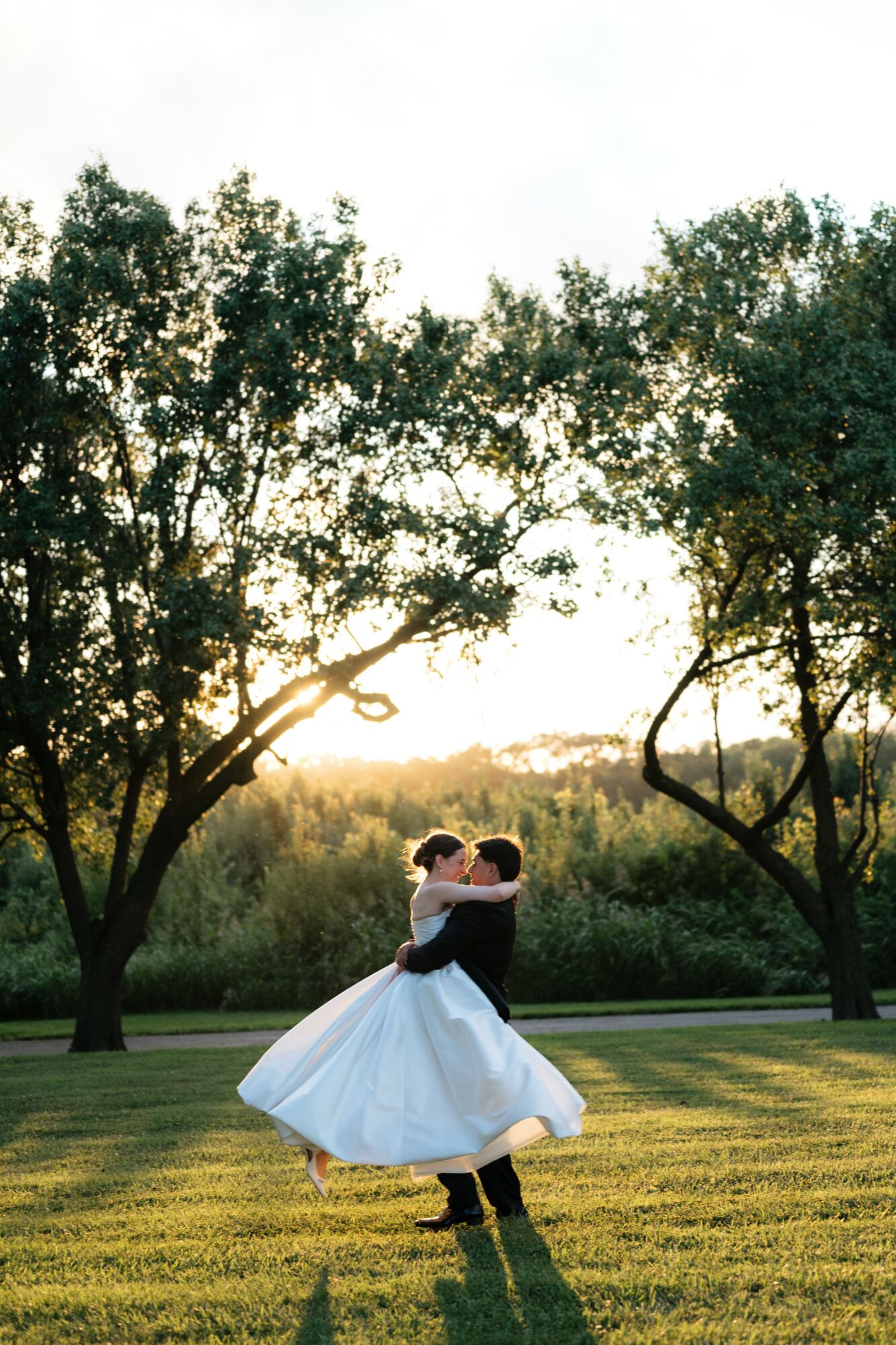 A couple dancing outdoors on grass, surrounded by trees, with sunlight in the background, embracing each other.