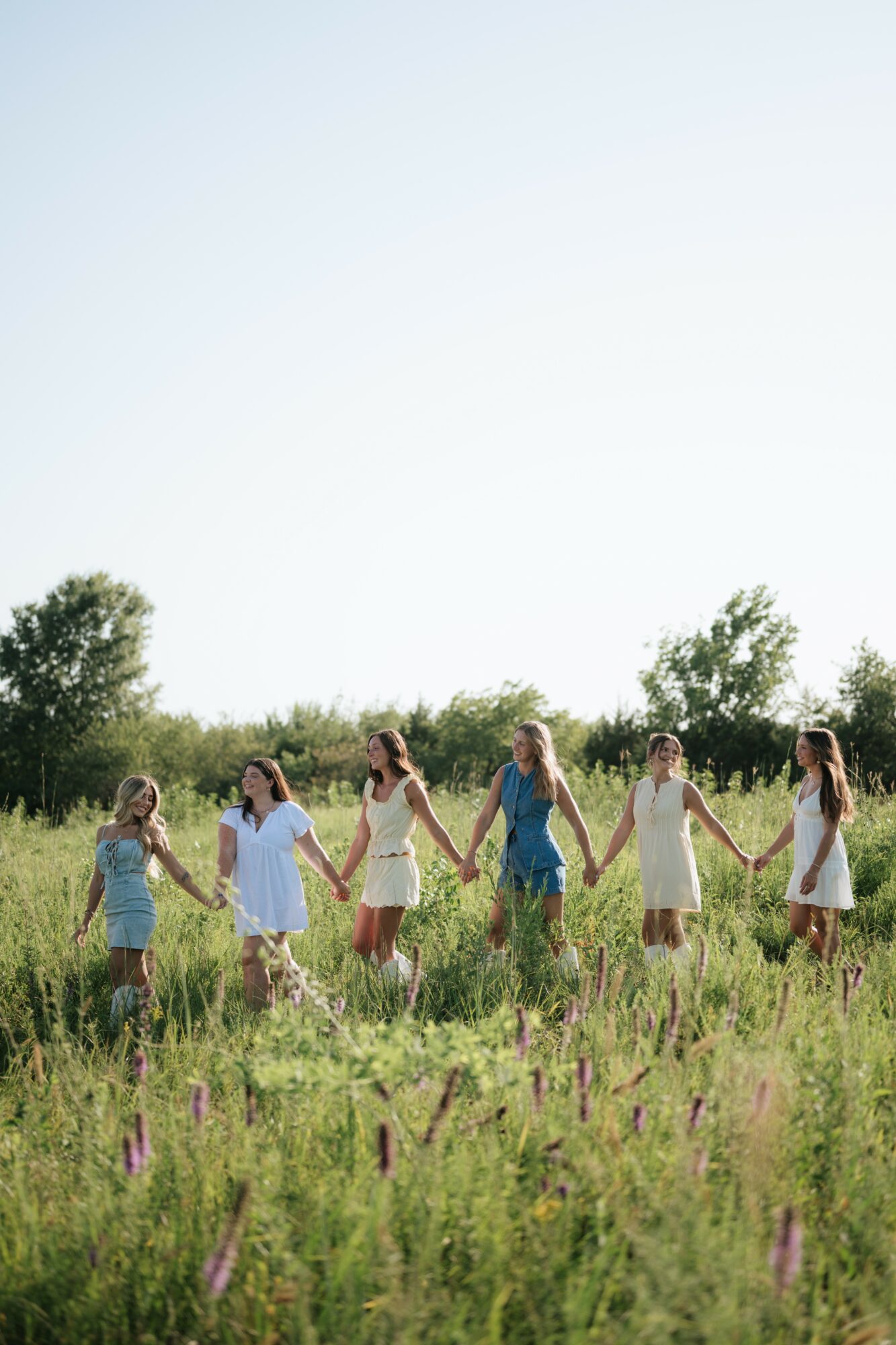 Group of children holding hands in a grassy field with trees and a clear sky in the background.