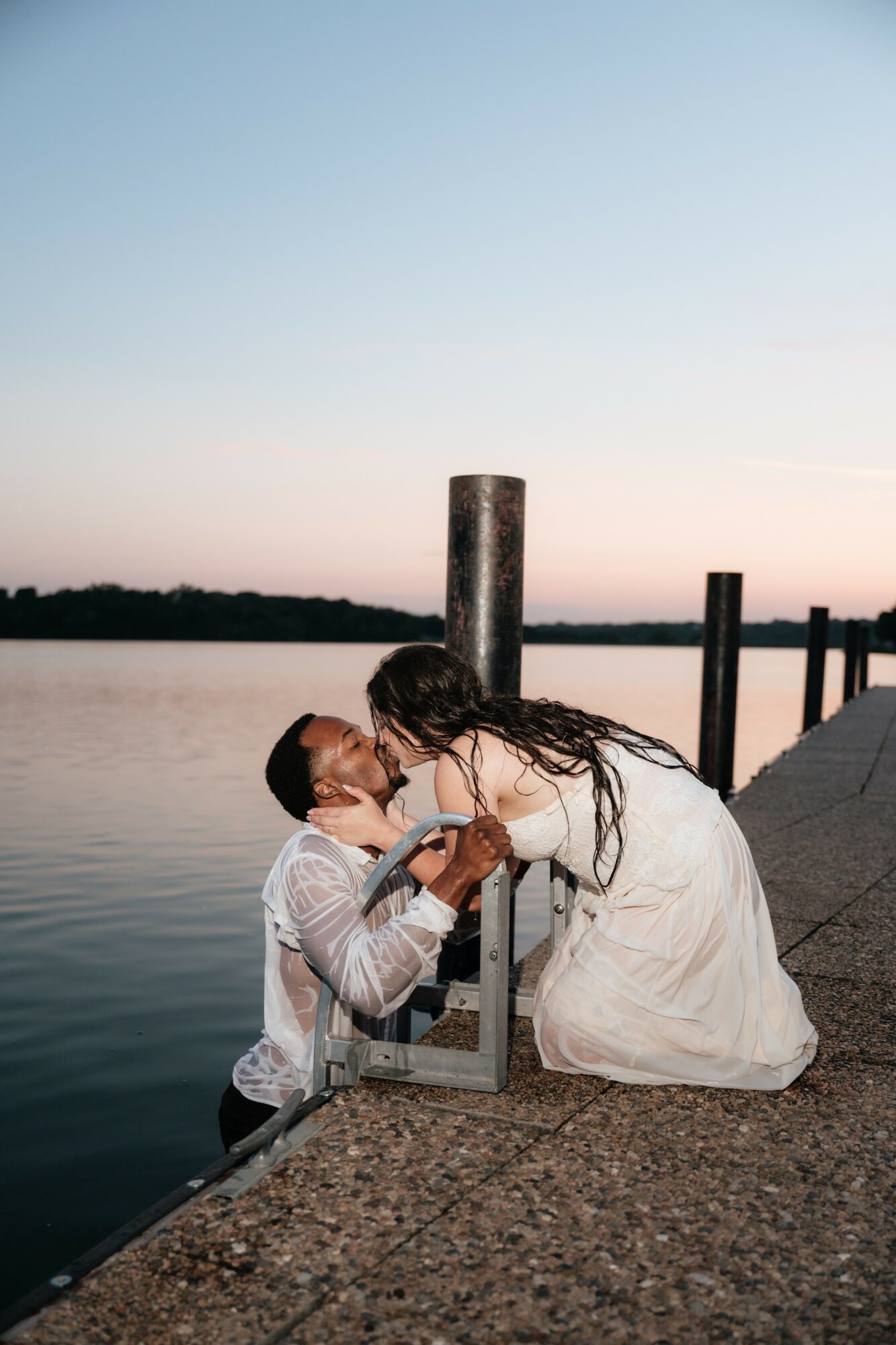 Two people on a dock by water, one kneeling and the other leaning forward, holding a framed photo, during sunset.