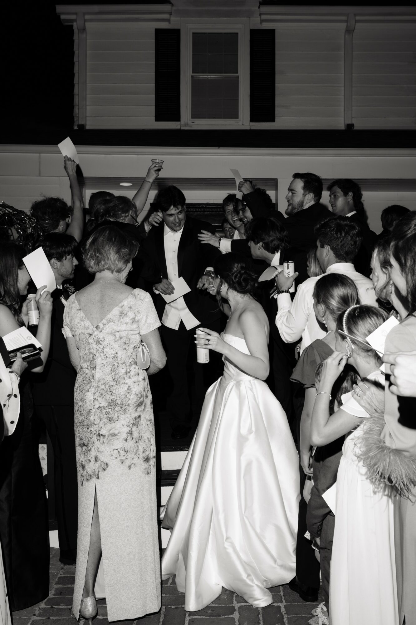 Group of people gathered indoors, some holding papers, with a woman in a long dress at the center.