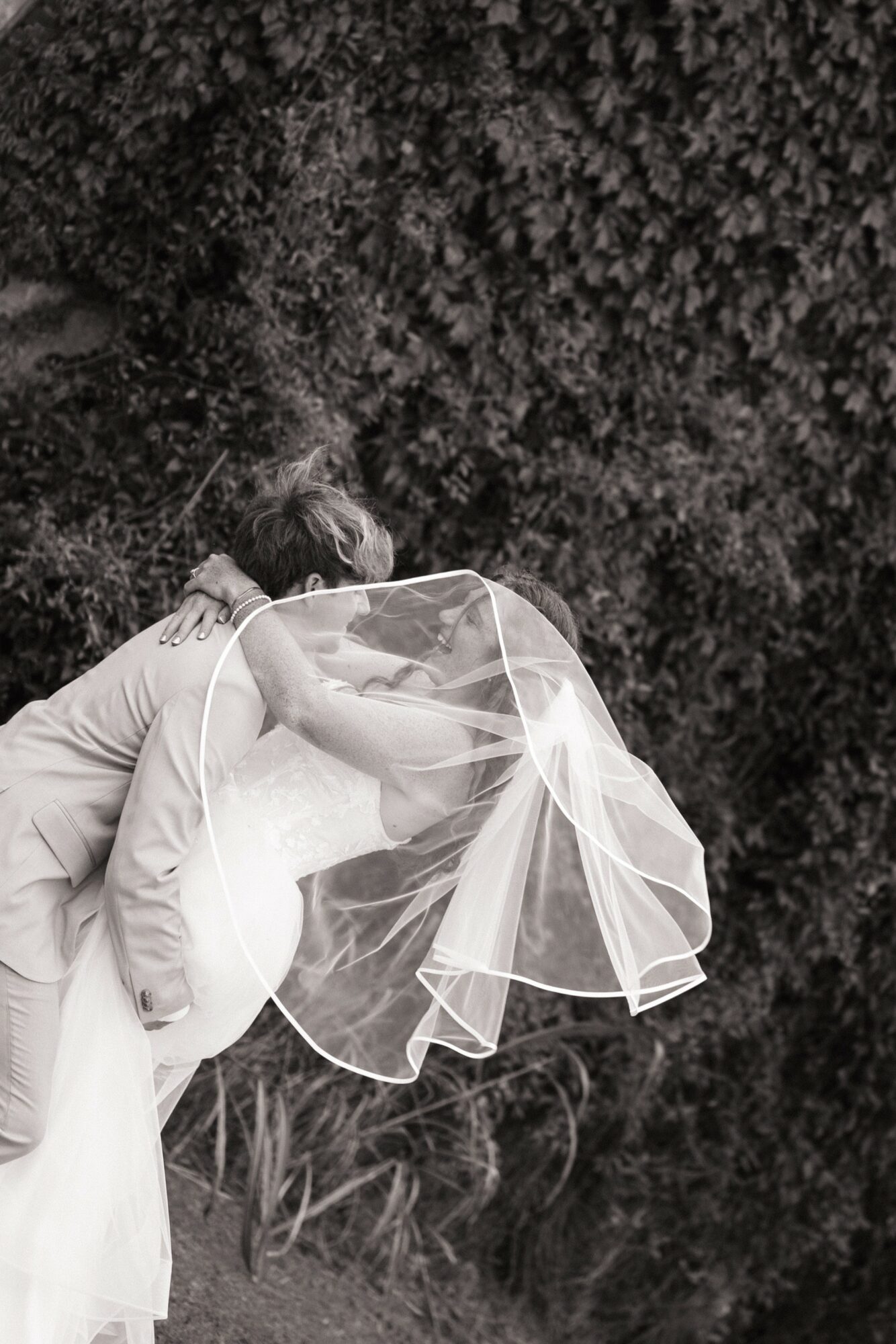 Person wearing a wedding veil and dress, holding a bouquet, standing outdoors near trees.