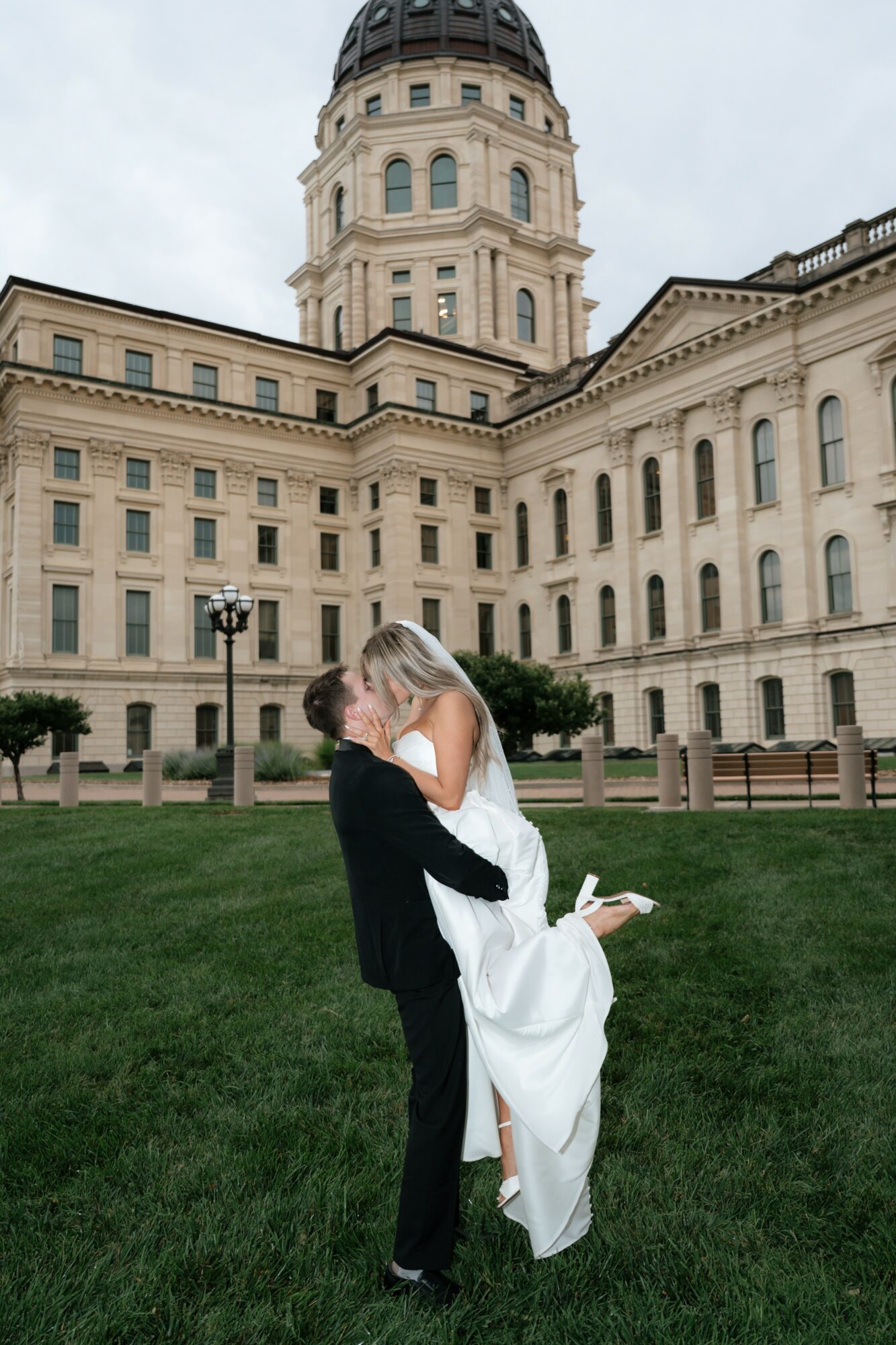 A couple in wedding attire embracing in front of a historic building with a tower, green grass in foreground.
