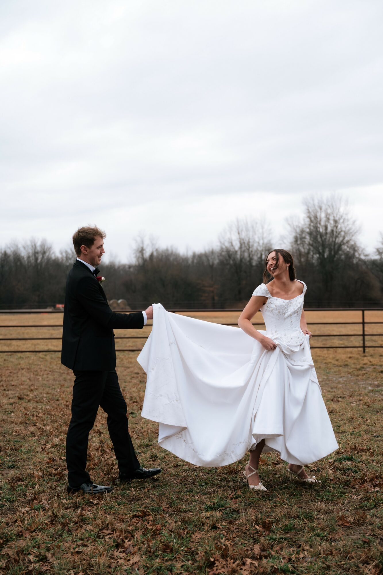 A couple in wedding attire holding hands outdoors, with a fence and trees in the background.