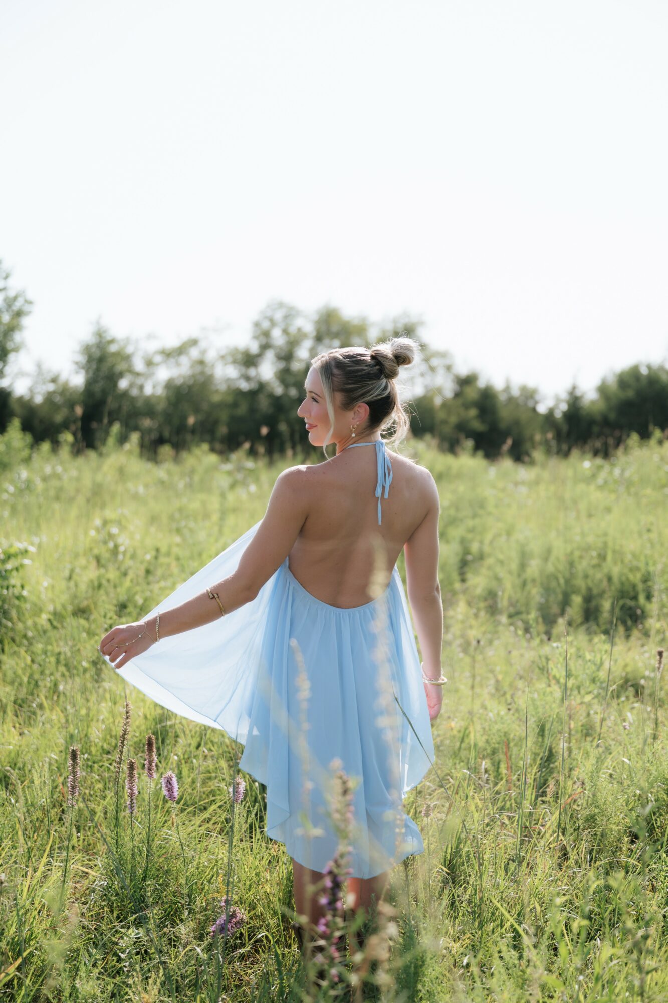 Woman in a light blue dress standing in a grassy field with trees in the background, facing sideways, holding the dress with one hand.