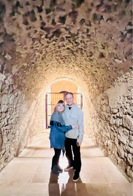Two people stand in a stone tunnel, holding hands and smiling, with a bright opening behind them.