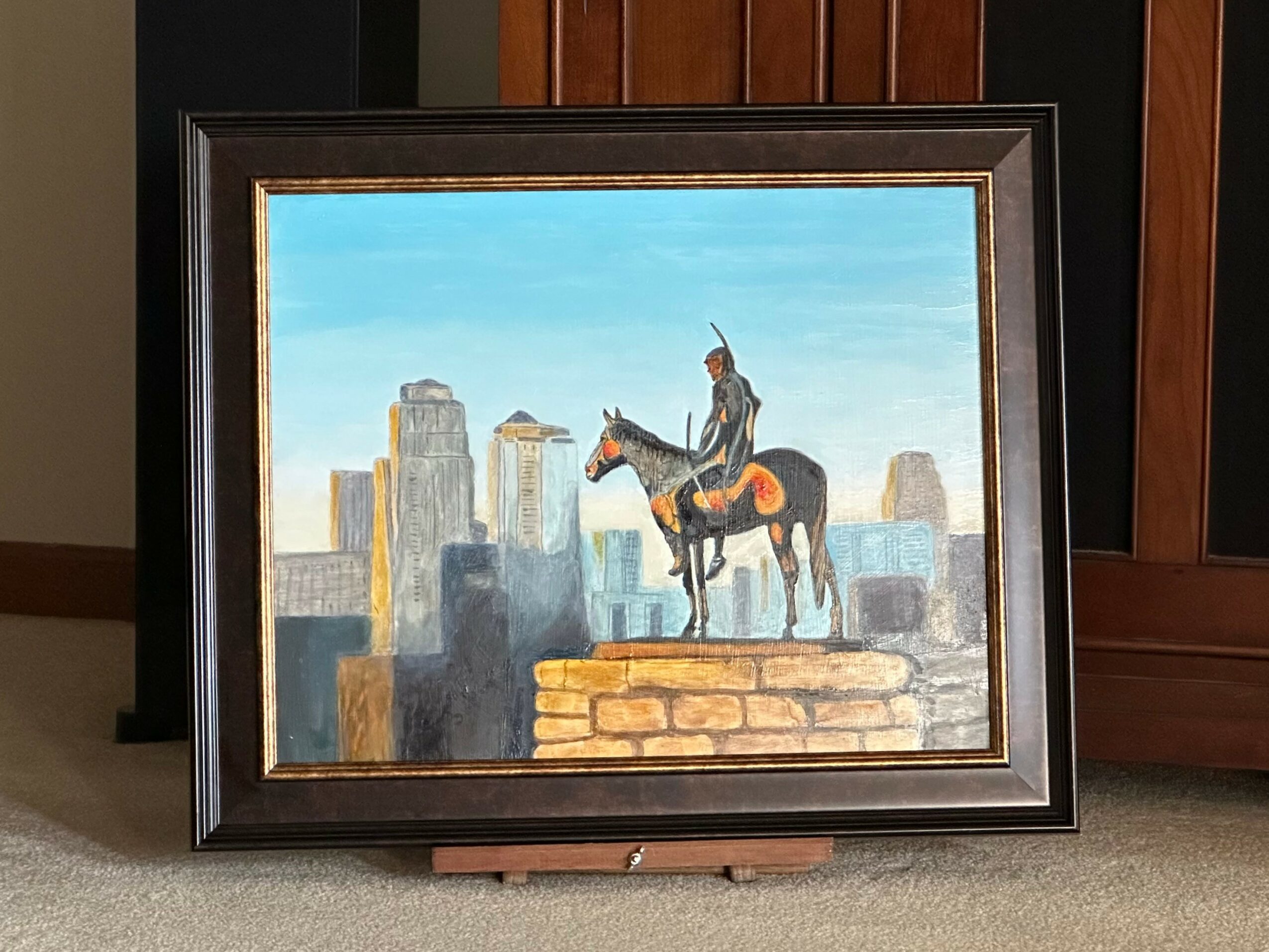 Framed painting of a person on a horse in front of a city skyline, with a blue sky background.