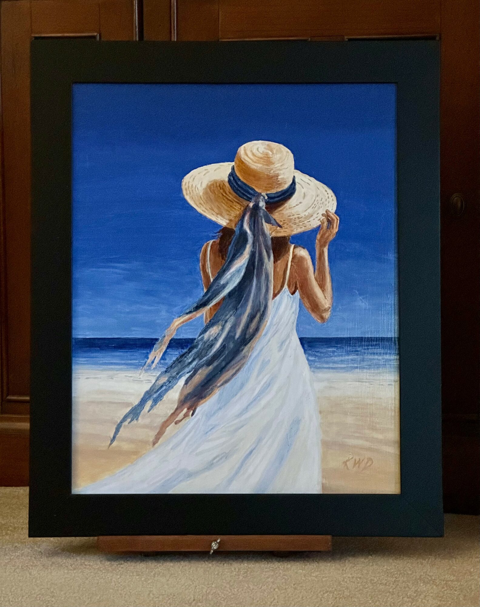 Painting of a woman in a white dress and large straw hat with a blue ribbon, standing on a beach with ocean in background.