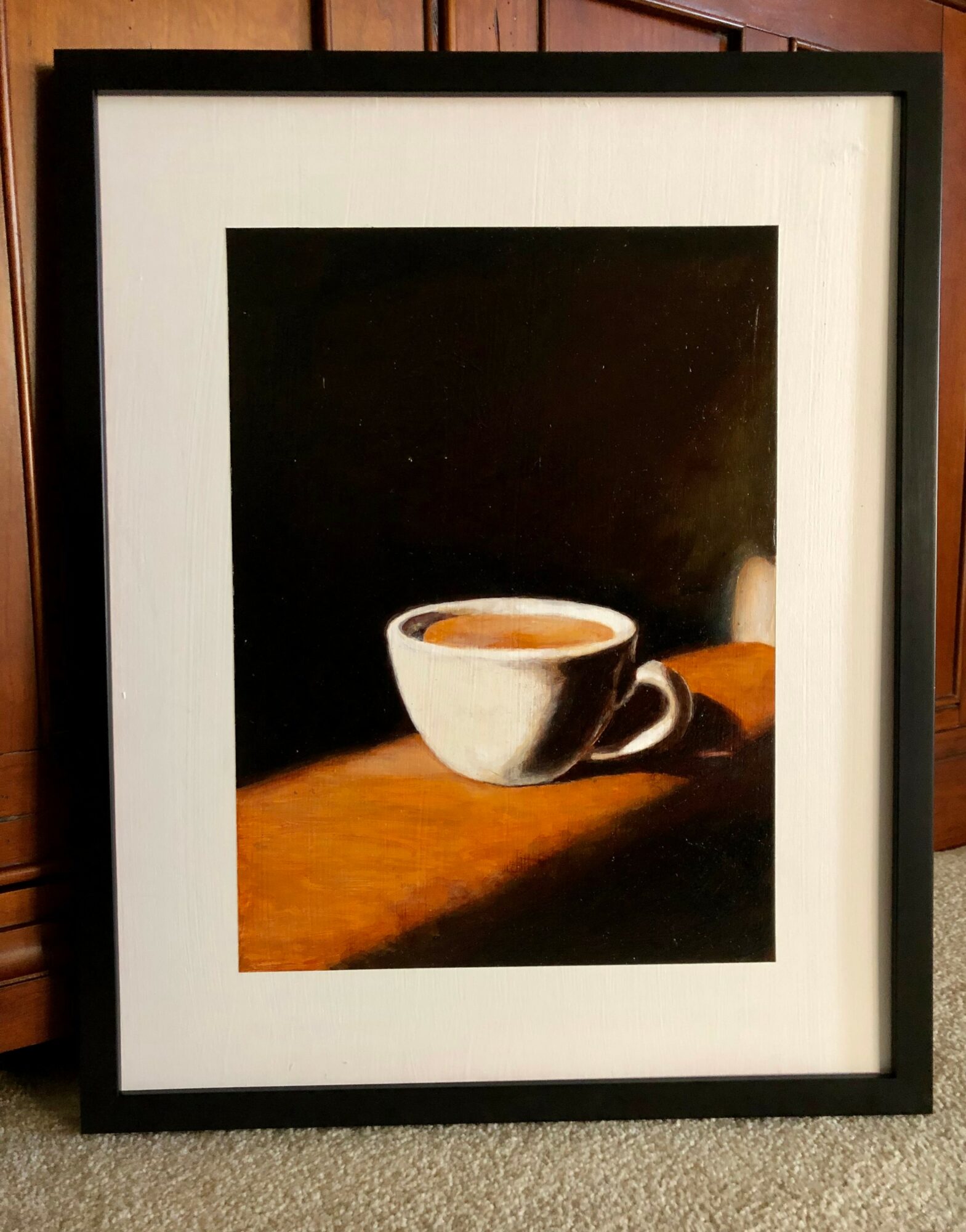 A framed painting of a white cup on a wooden surface with a dark background.