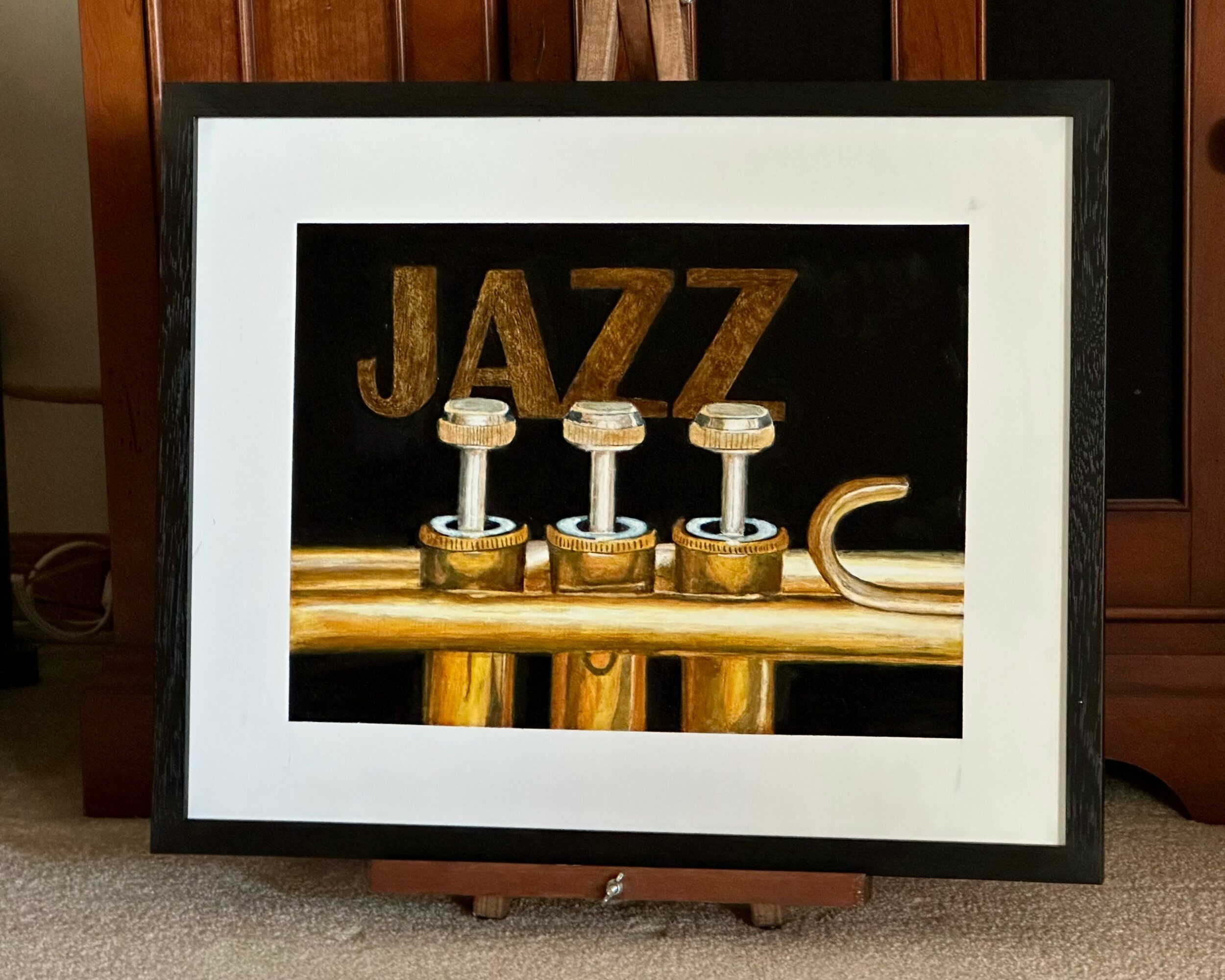 Framed artwork of a trumpet with the word JAZZ in gold letters above it, displayed on a wall or stand.