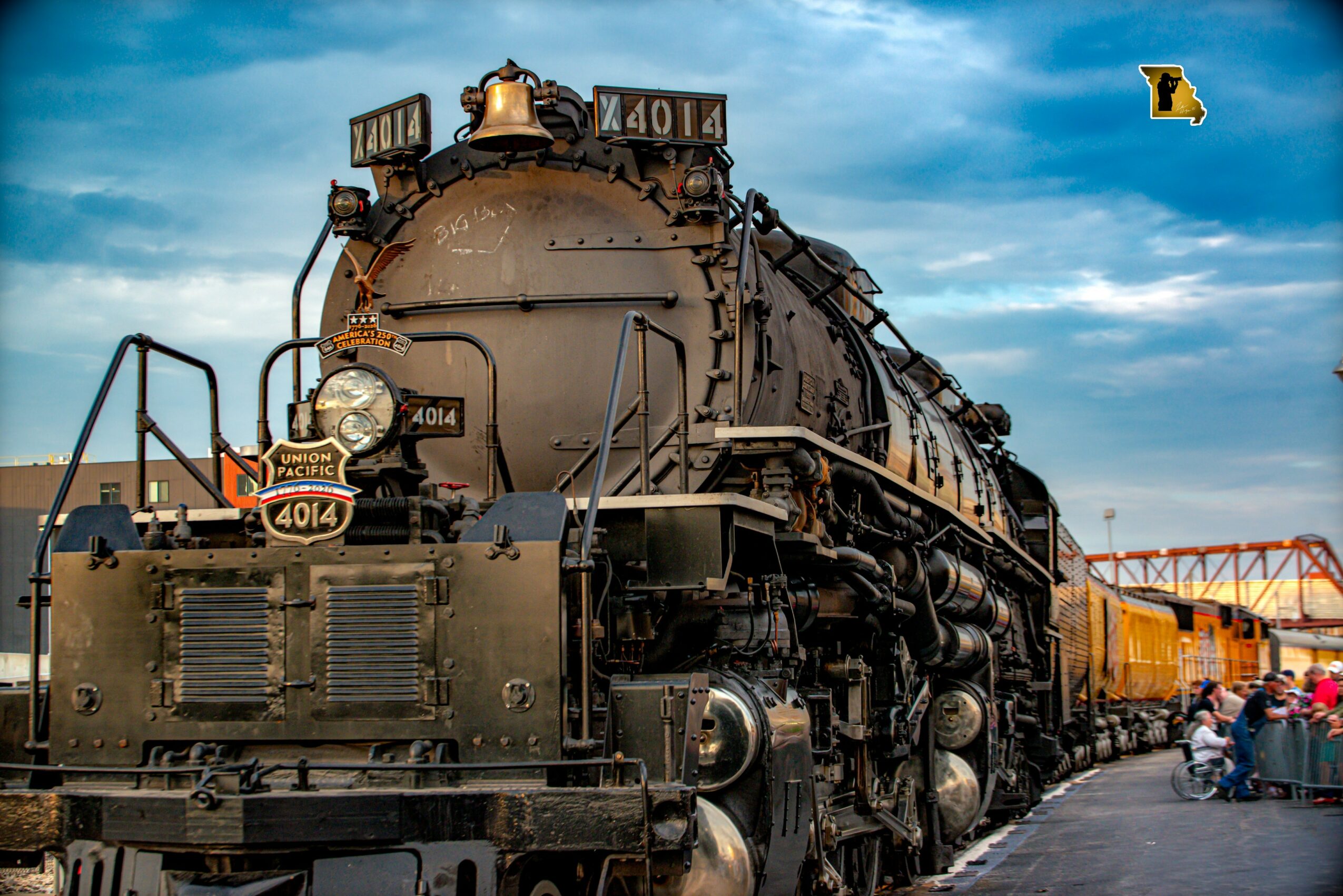 Steam locomotive train with yellow cars on the right, people nearby, blue sky with clouds, and a bridge in the background.