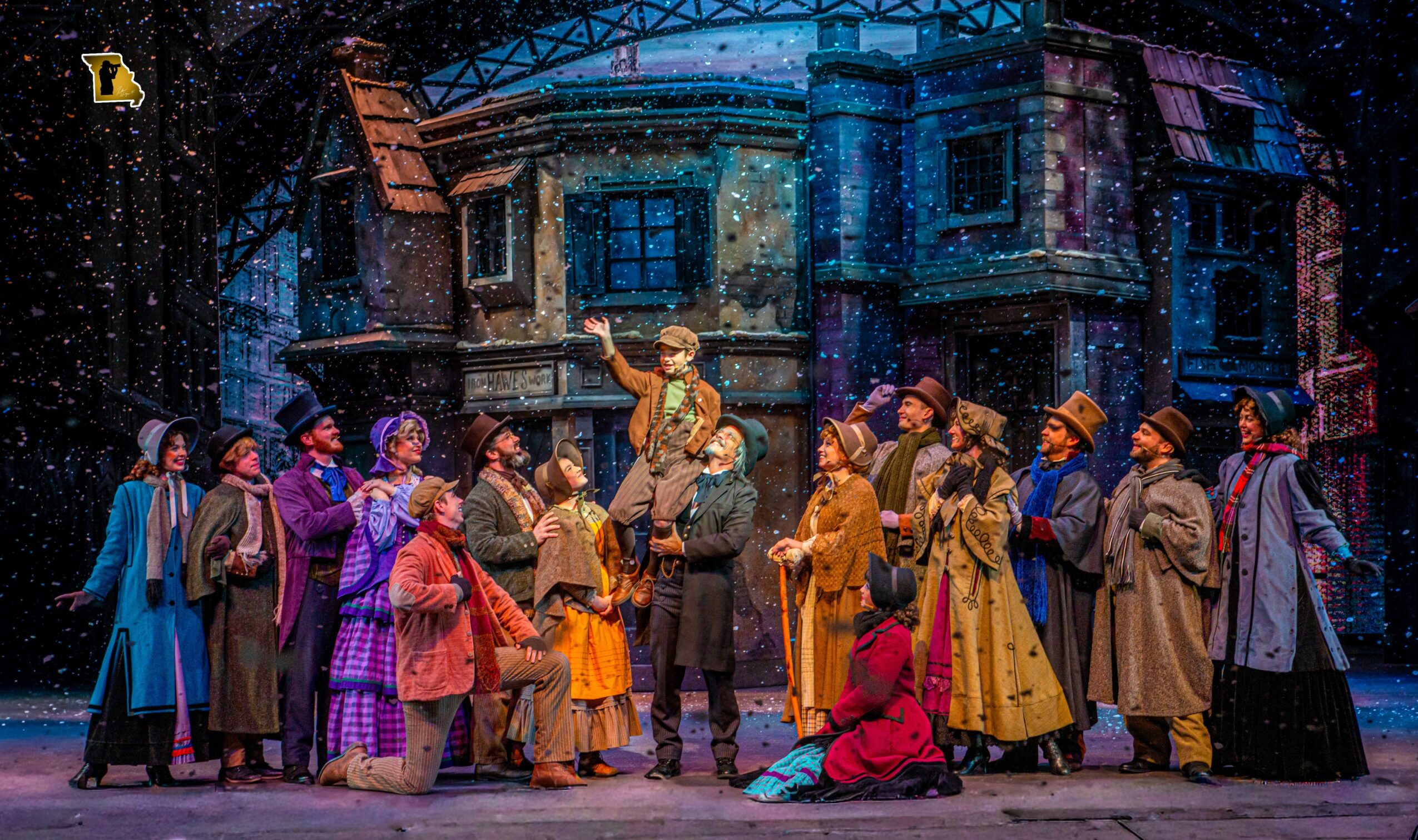 Group of performers in colorful costumes on stage with a building backdrop, engaged in a theatrical scene.