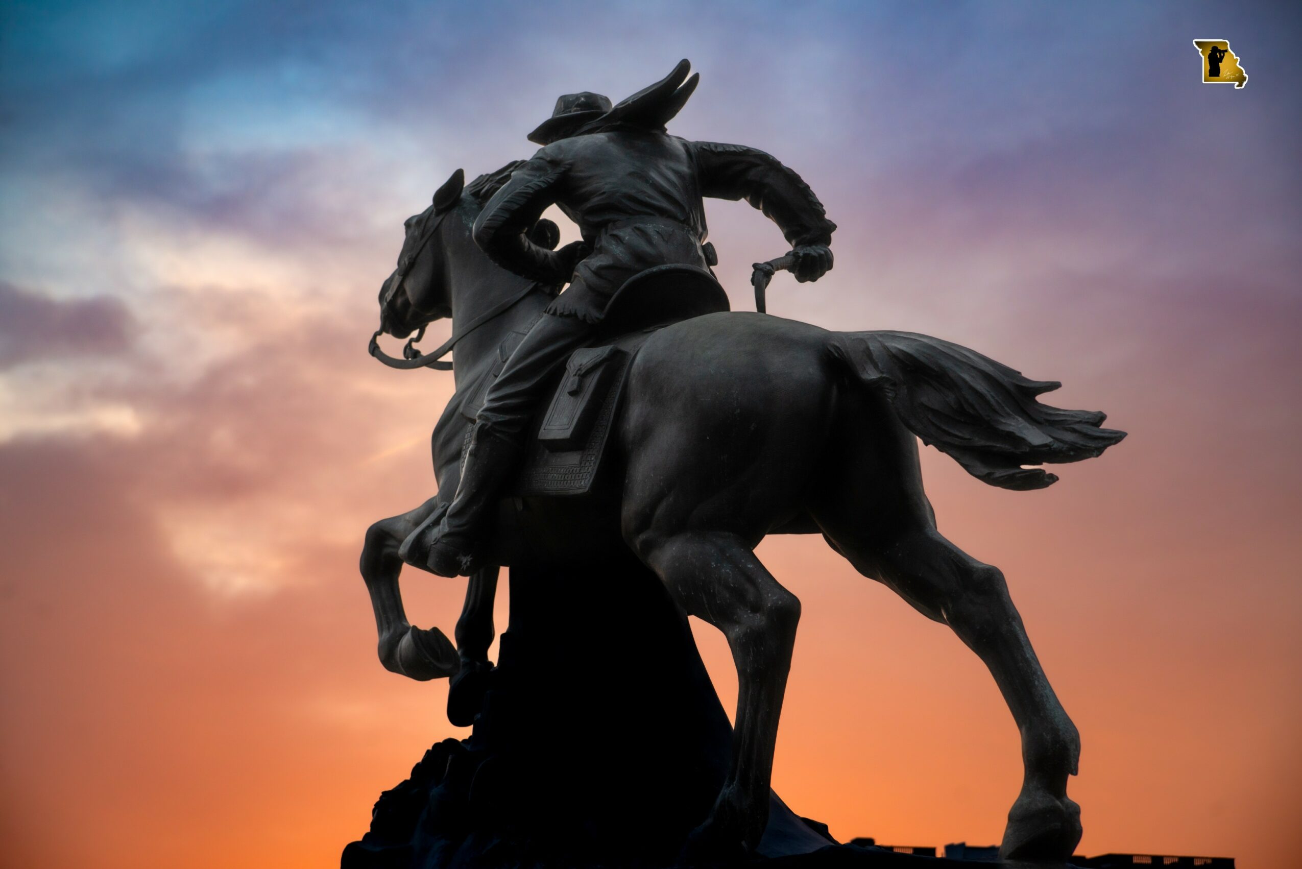 Statue of a person riding a horse against a colorful sunset sky, with clouds and a silhouette of the ground.