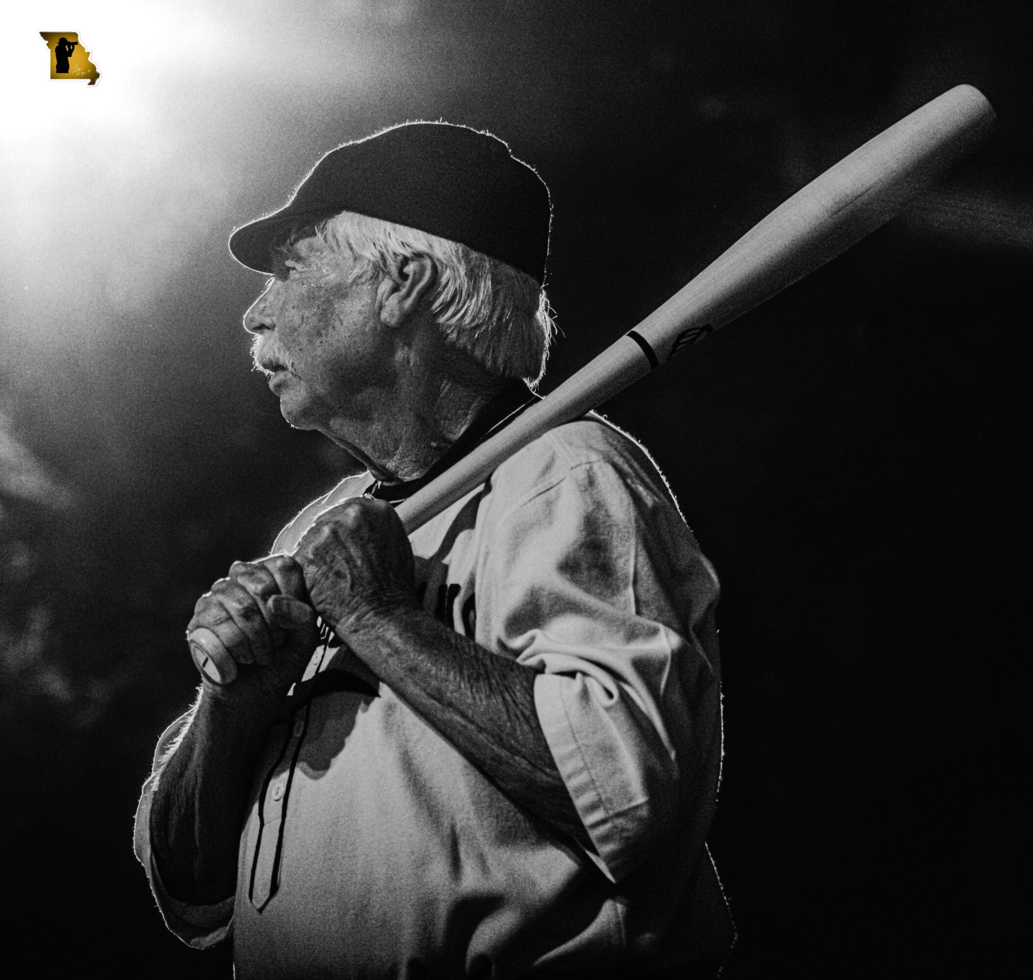 Elderly man with a baseball bat, wearing a cap, in black and white, looking to the side.