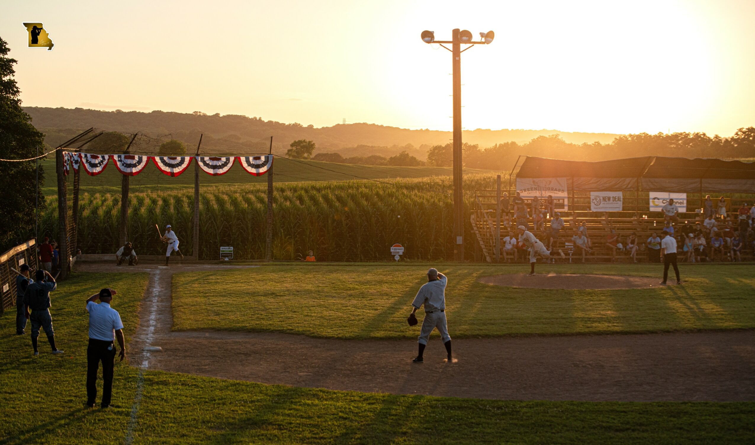 Baseball game at sunset with players, spectators, and decorated flags, field and power pole visible.