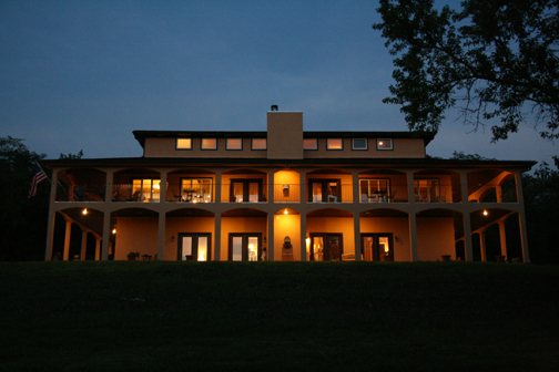 Large house with multiple balconies illuminated at night, surrounded by trees and a dark sky.
