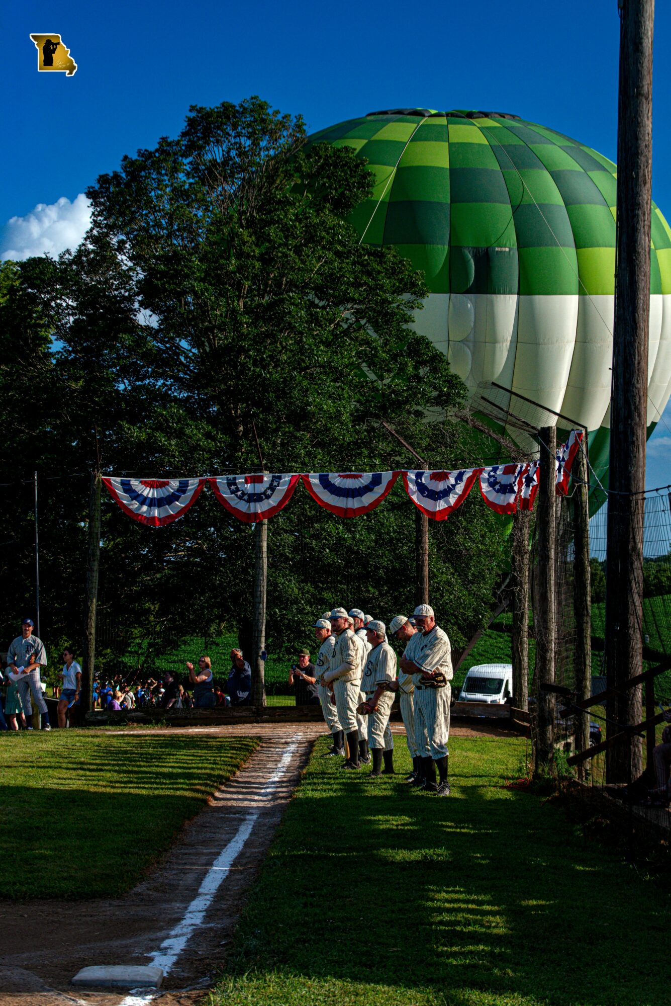 People gather outdoors near a hot air balloon with green and white sections, decorated with patriotic bunting, under a blue sky.