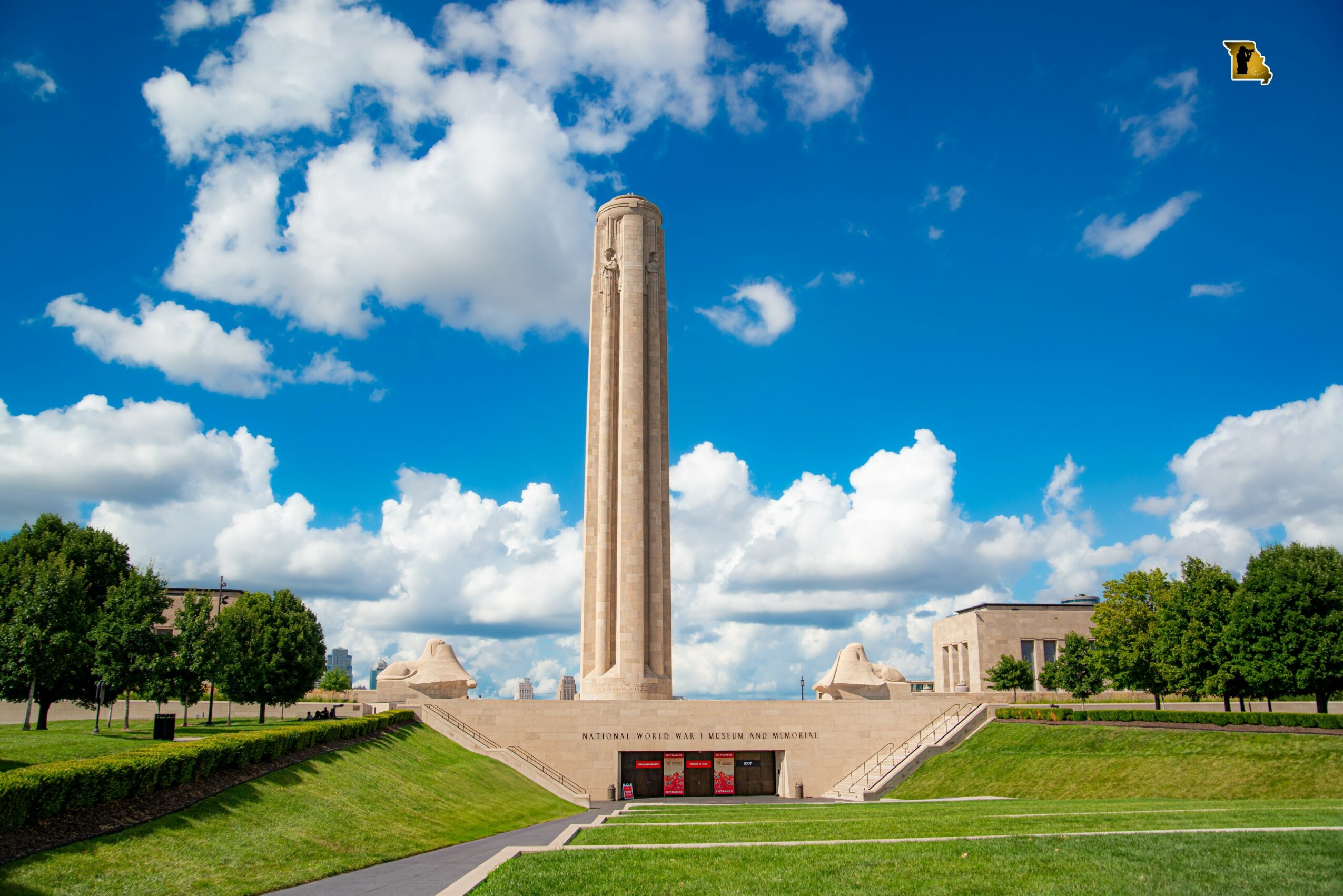 Tall monument with a blue sky and clouds, surrounded by green trees and a grassy area.
