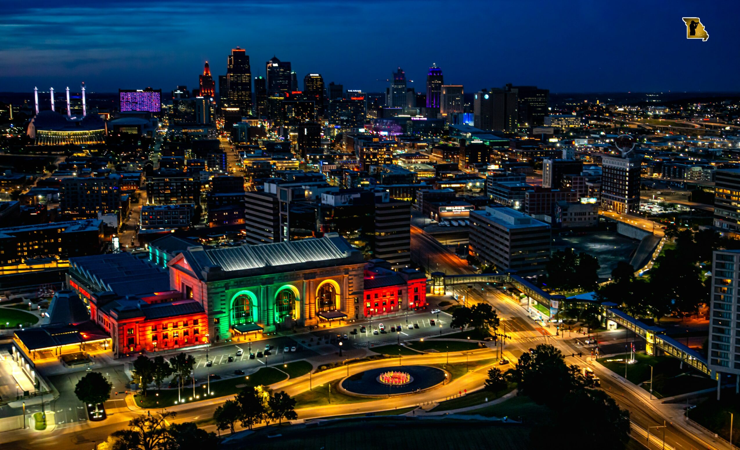 Nighttime cityscape with illuminated buildings and a historic train station decorated with green, red, and yellow lights, and a roundabout in foreground.