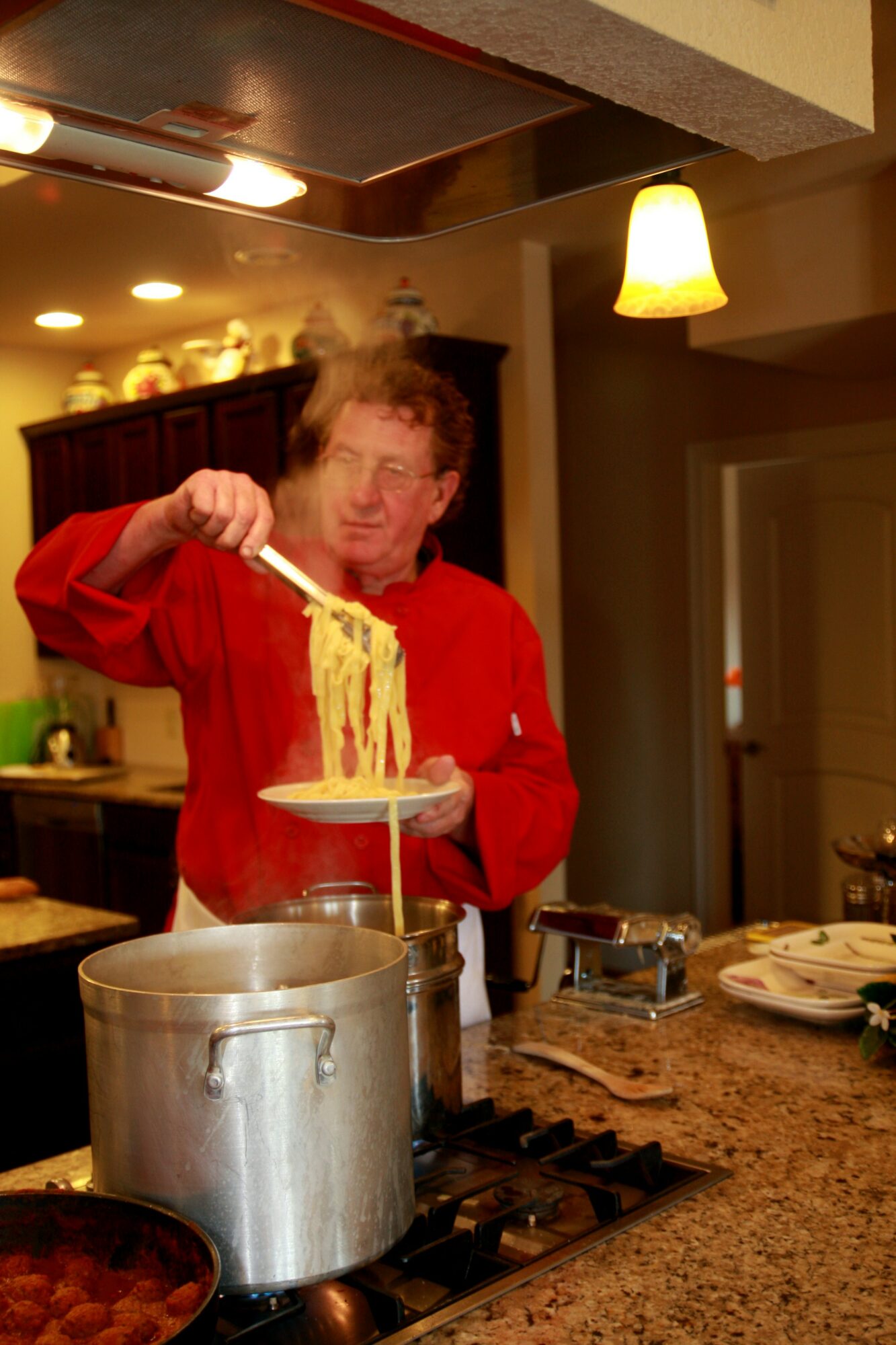 Person in red shirt lifting a plate of steaming noodles over a pot in a kitchen, with warm lighting and cabinets in background.