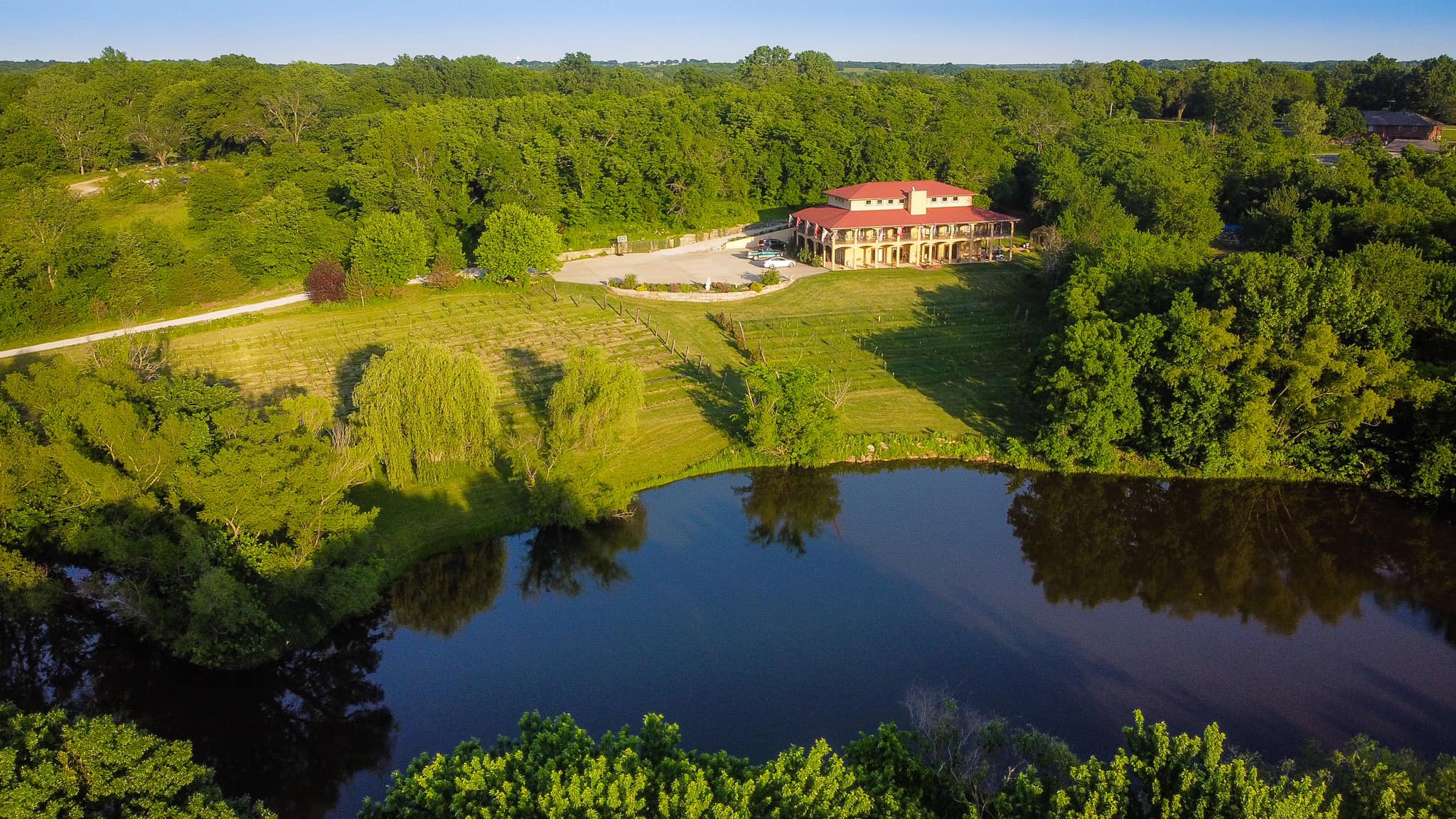 A large house with a red roof is situated on a hill surrounded by trees, overlooking a pond with reflections.