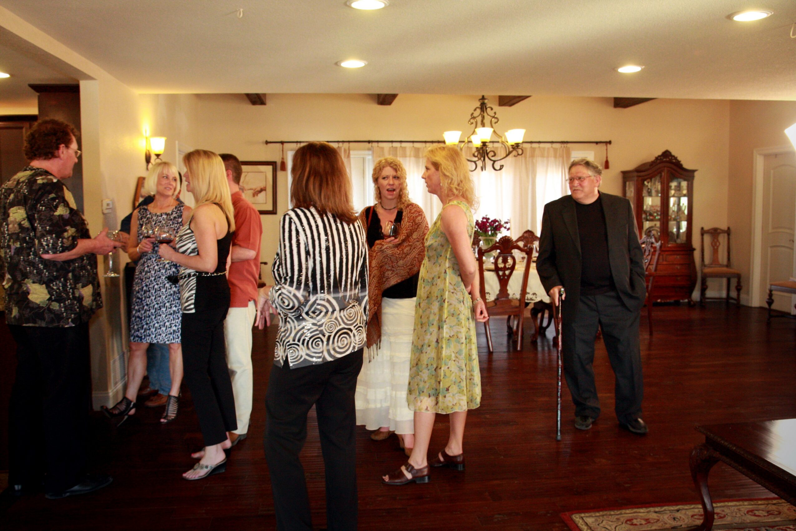 Group of people socializing indoors, some holding drinks, in a room with wooden floors and elegant furniture.