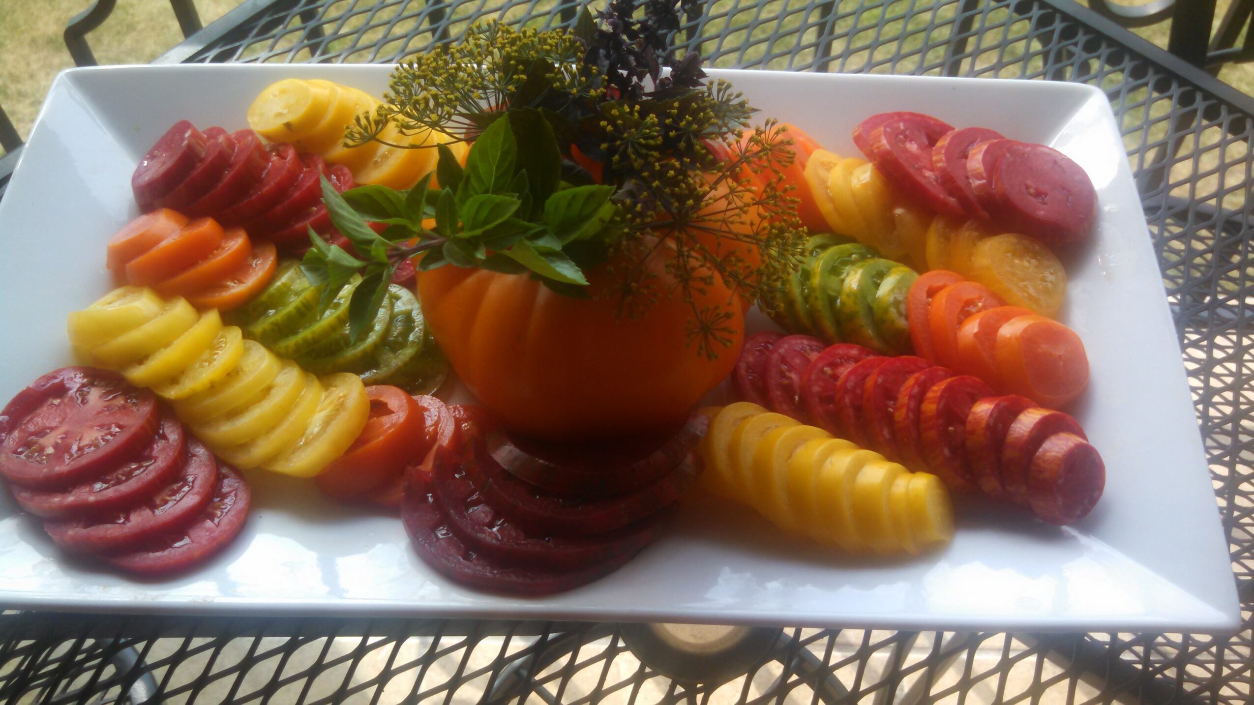 Colorful spiral-cut vegetables arranged around a central orange flower in a vase on a white rectangular plate.