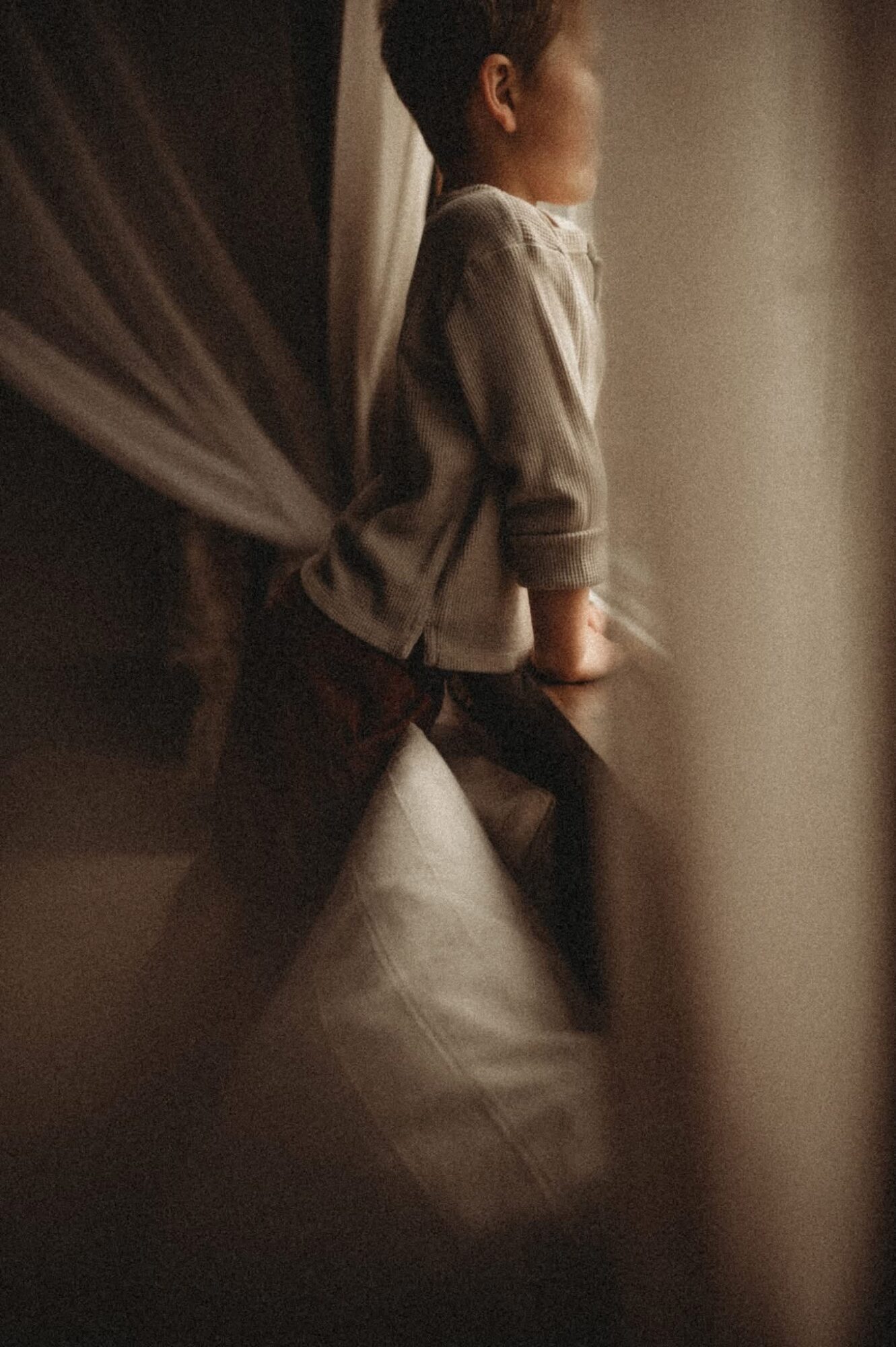 Child standing by a window, looking outside, with soft lighting and curtains nearby.