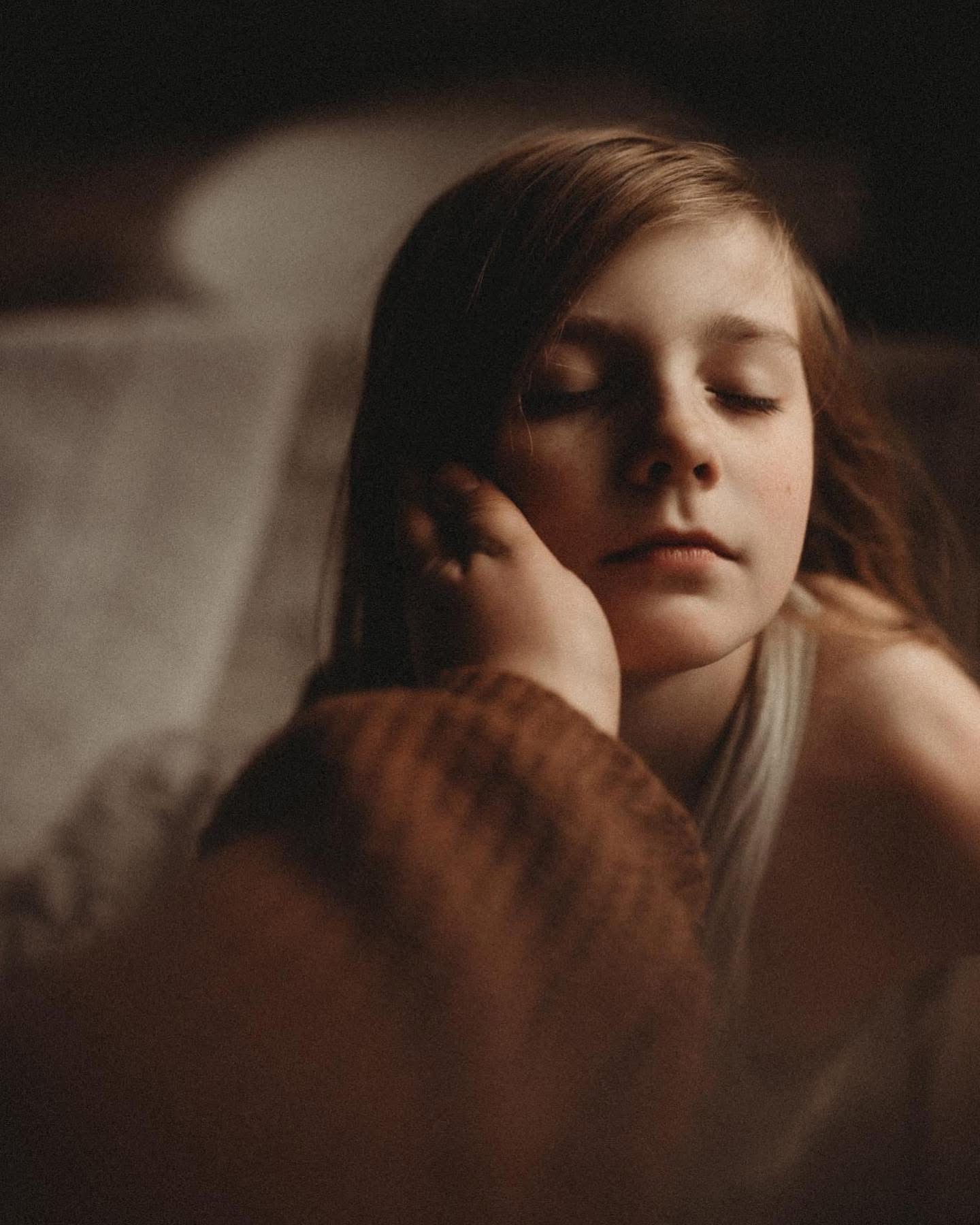 Young girl with closed eyes resting her face on her hand, in a dimly lit setting.