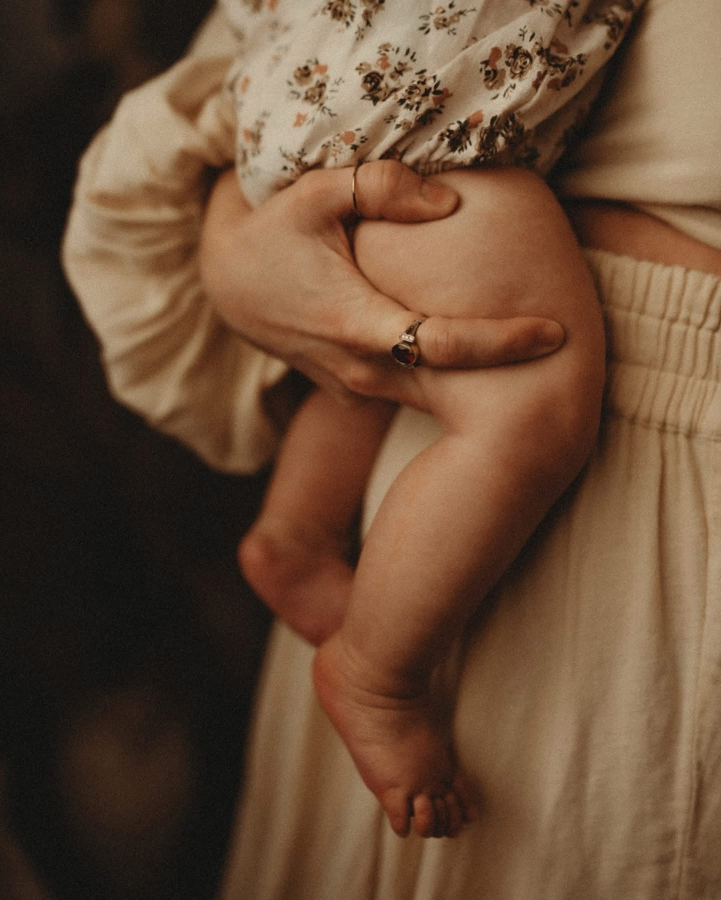 Person holding a child's leg, with the child's foot hanging down, wearing a floral dress and beige clothing.