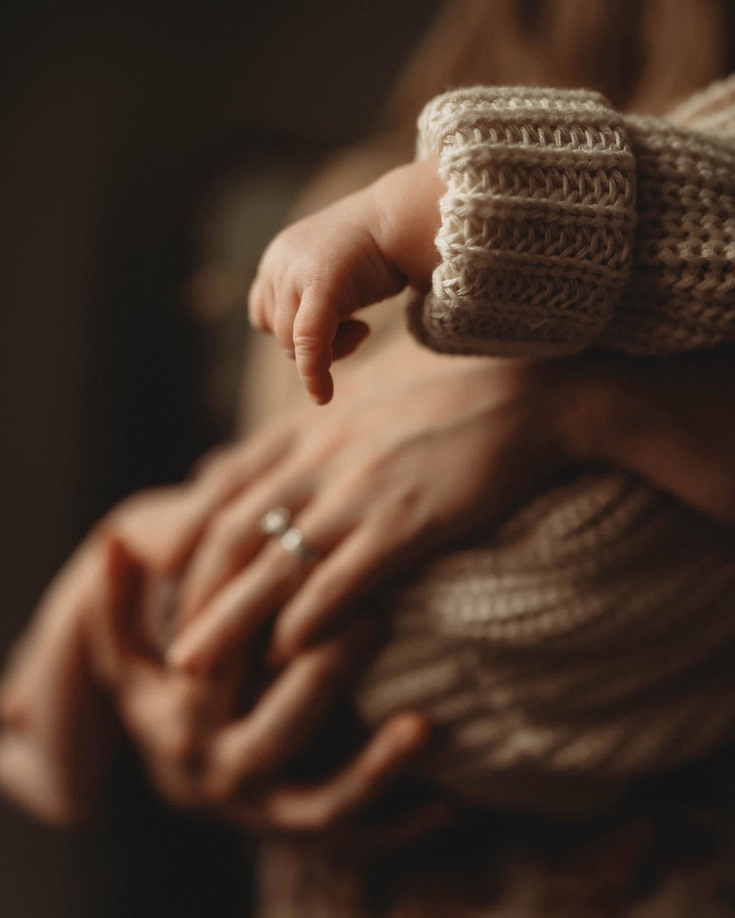 Close-up of a person's hand holding a baby's hand, both wearing knitted clothing, warm indoor setting.