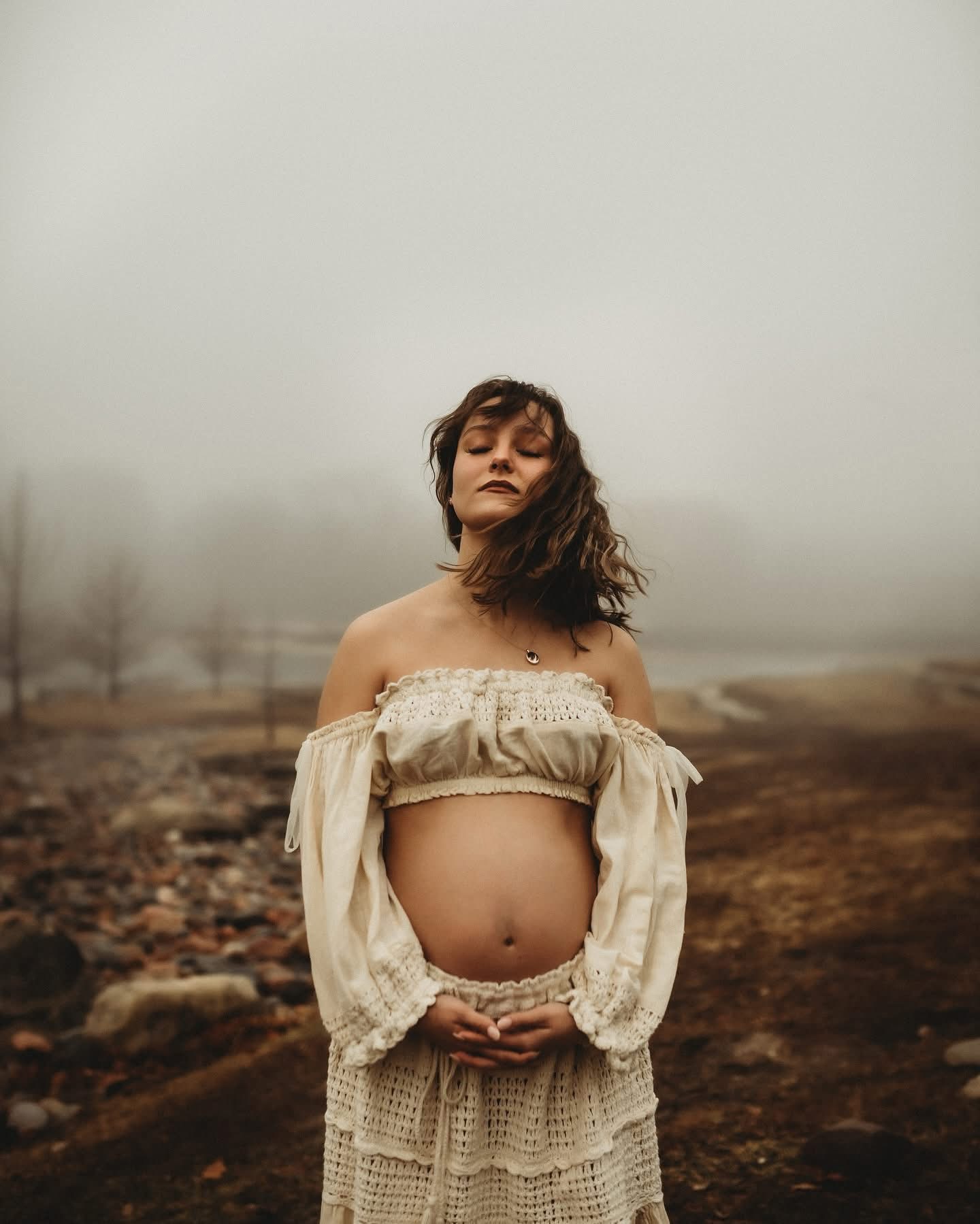 Pregnant woman standing outdoors with hands on belly, wearing off-shoulder dress, foggy landscape background.