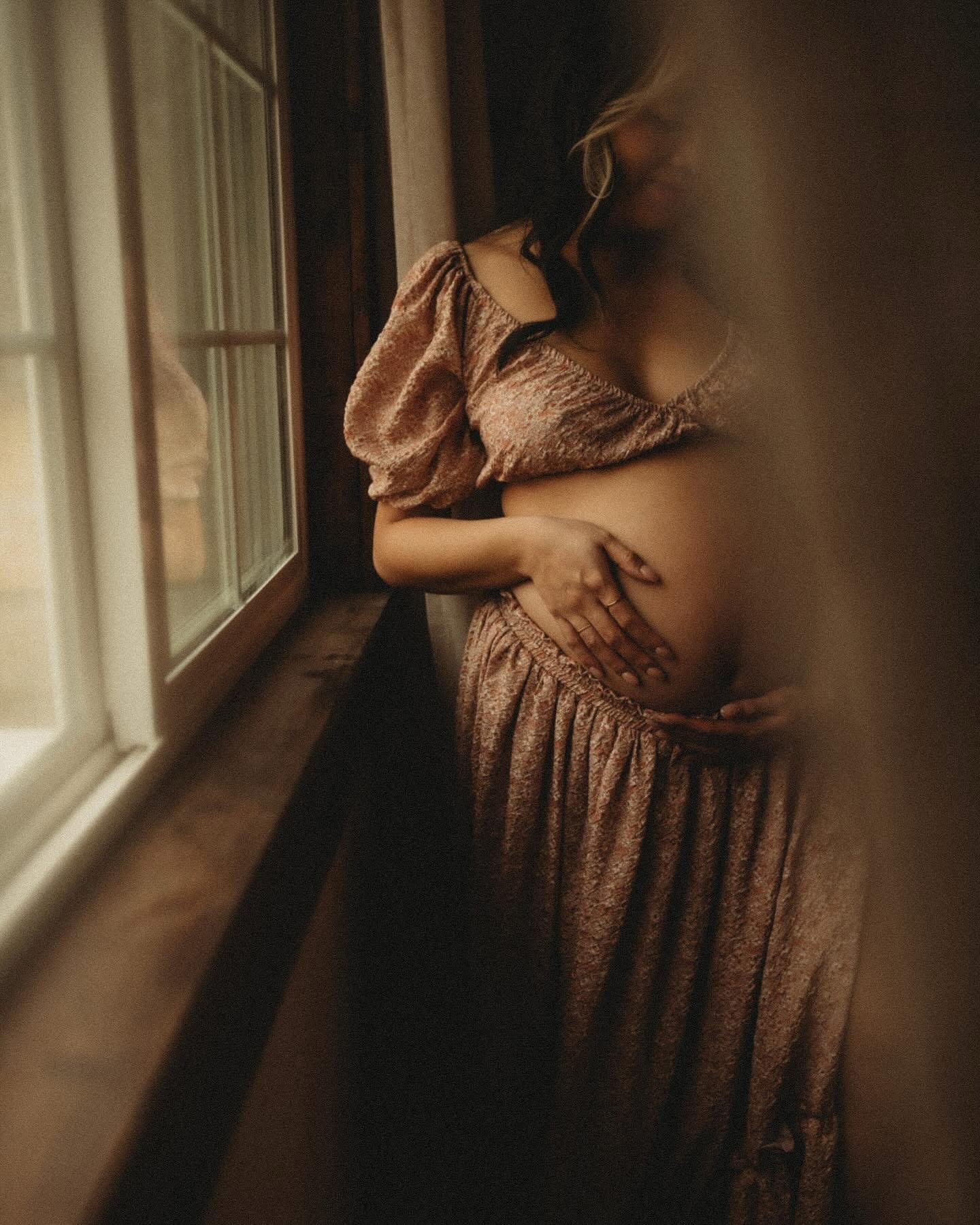Pregnant woman standing by window, wearing a flowing dress, with her hand on her belly, soft lighting, blurred background.