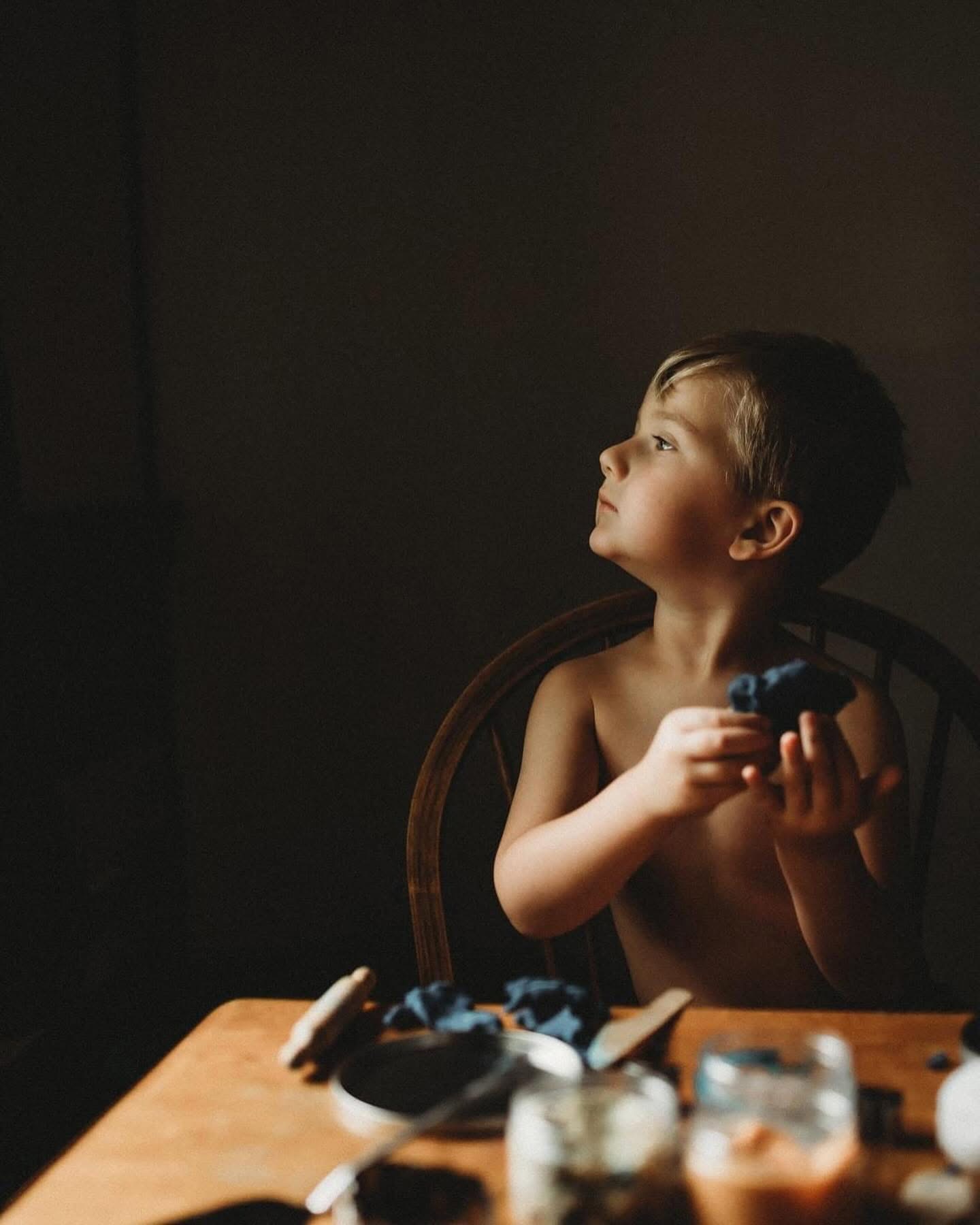 A young boy with short dark hair sits shirtless at a wooden table, holding a toy, looking upward with a thoughtful expression.