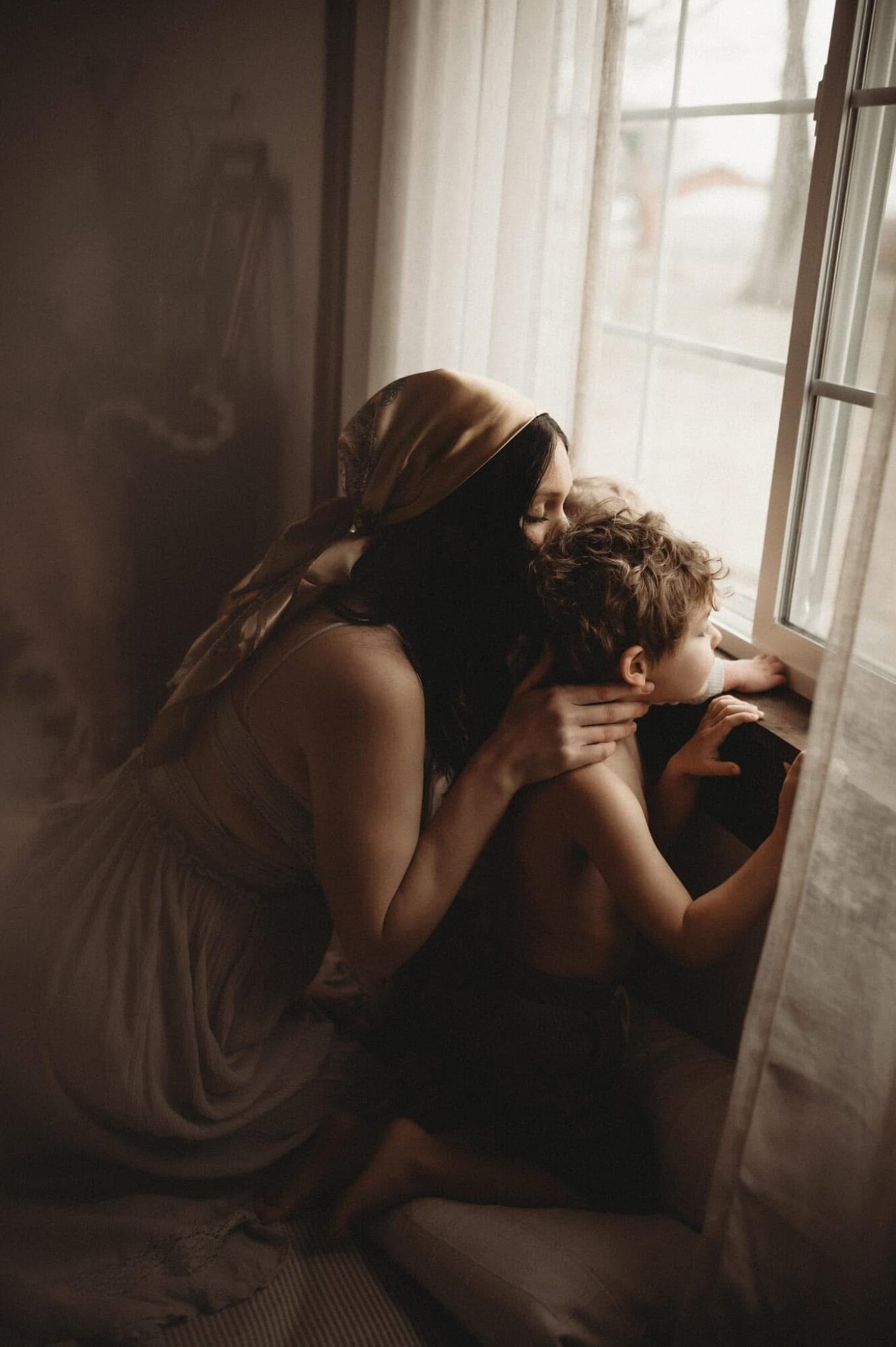 Woman and child looking out a window, sitting on a bed near curtains, with sunlight coming through the window.
