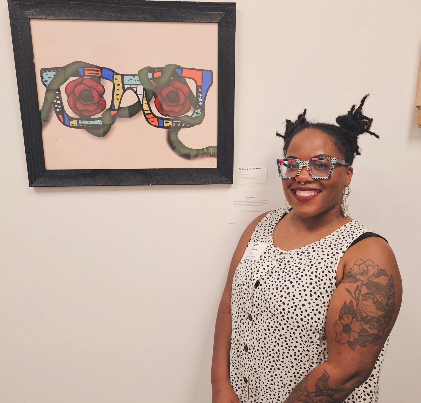 Smiling woman with glasses and tattoos stands next to colorful framed artwork of two cameras on a white wall.