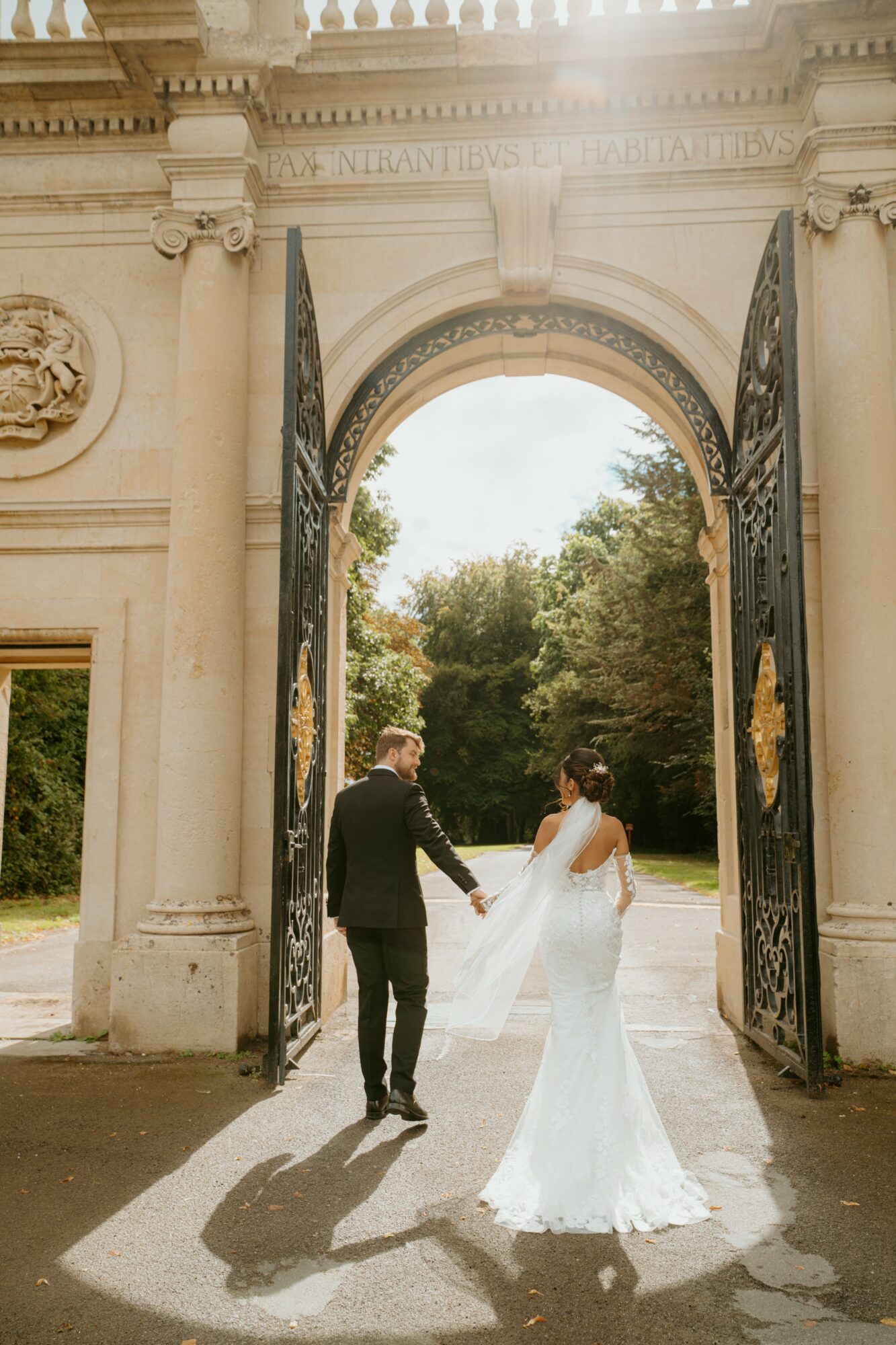 A couple in wedding attire holding hands under an ornate archway, with trees and sky in the background.