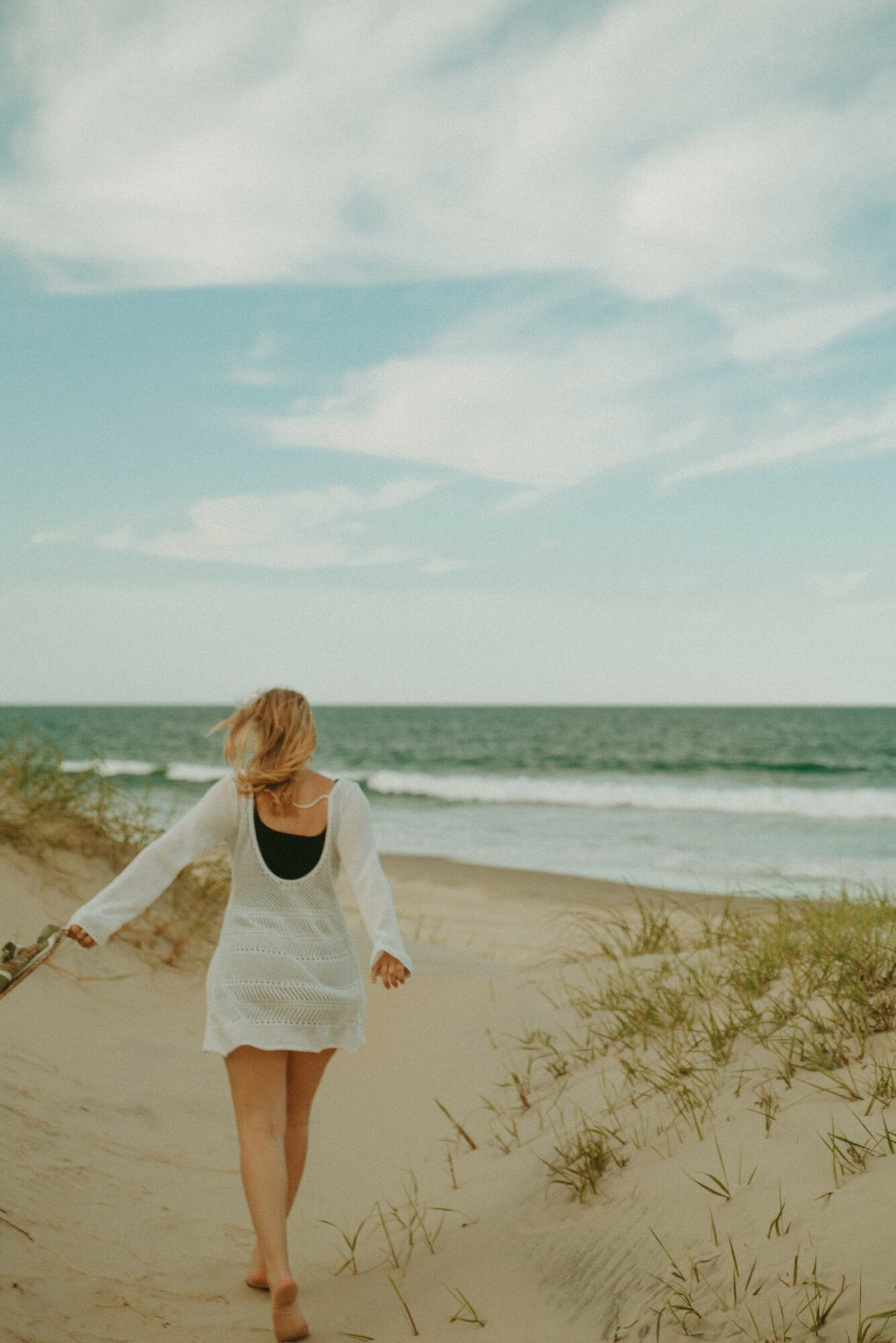 Woman walking barefoot on sandy beach with ocean and sky in background, holding a branch, hair blowing in wind.