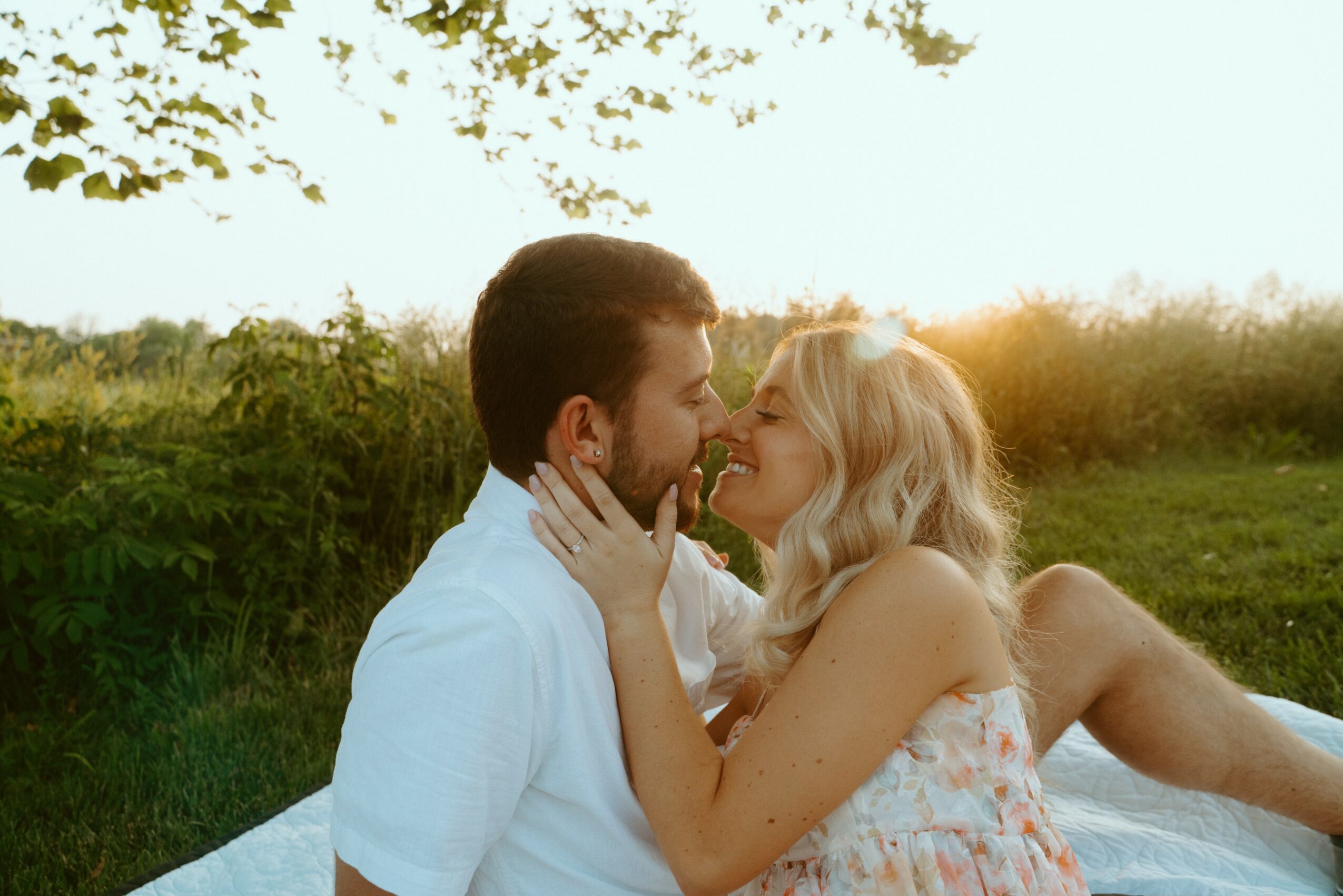 A couple lying on grass outdoors, facing each other, smiling, with sunlight in the background.