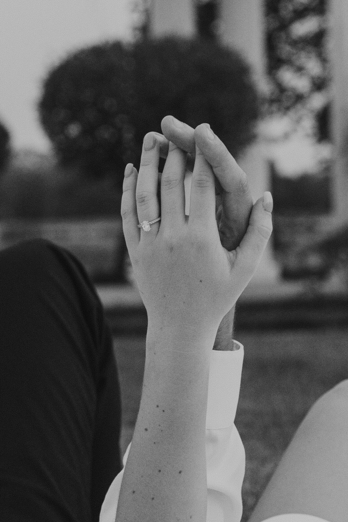Two hands with wedding rings gently touching, outdoors with blurred trees and bushes in background.
