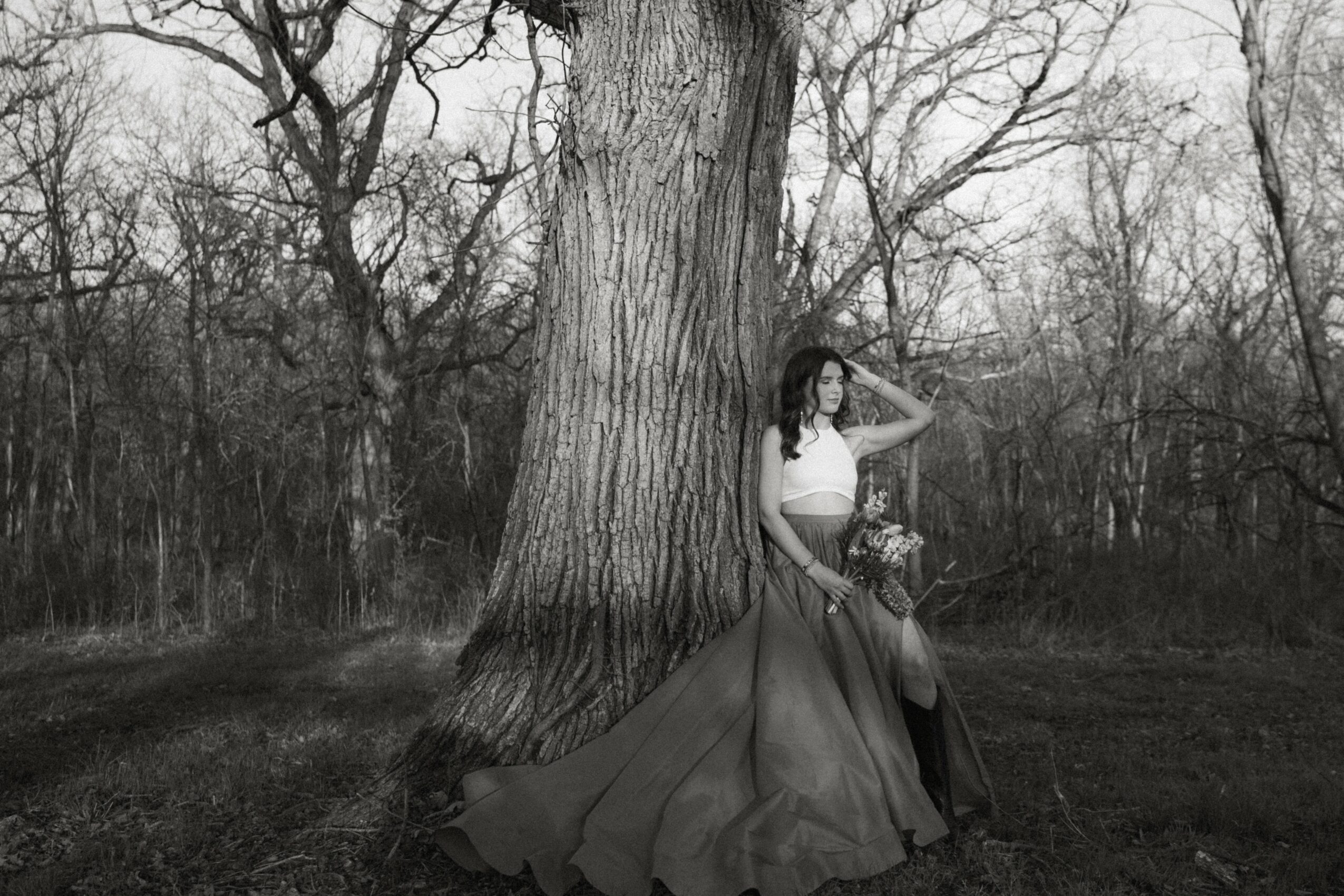 Woman sitting on a chair outdoors near a large tree, holding a bouquet, in a black-and-white photo.