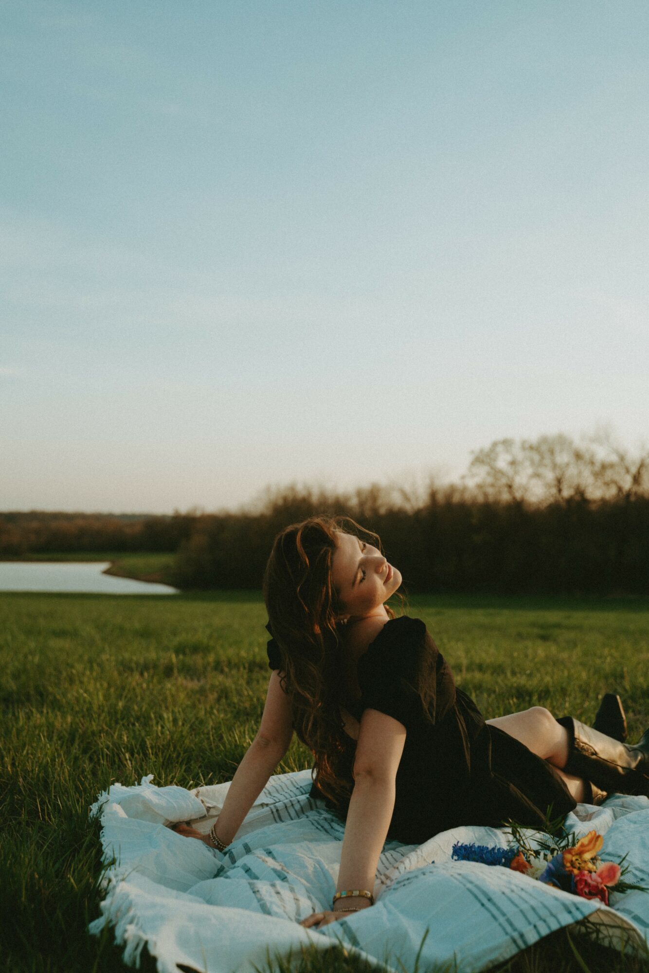 Person sitting on grass near water, looking up, with a landscape and sky in the background.