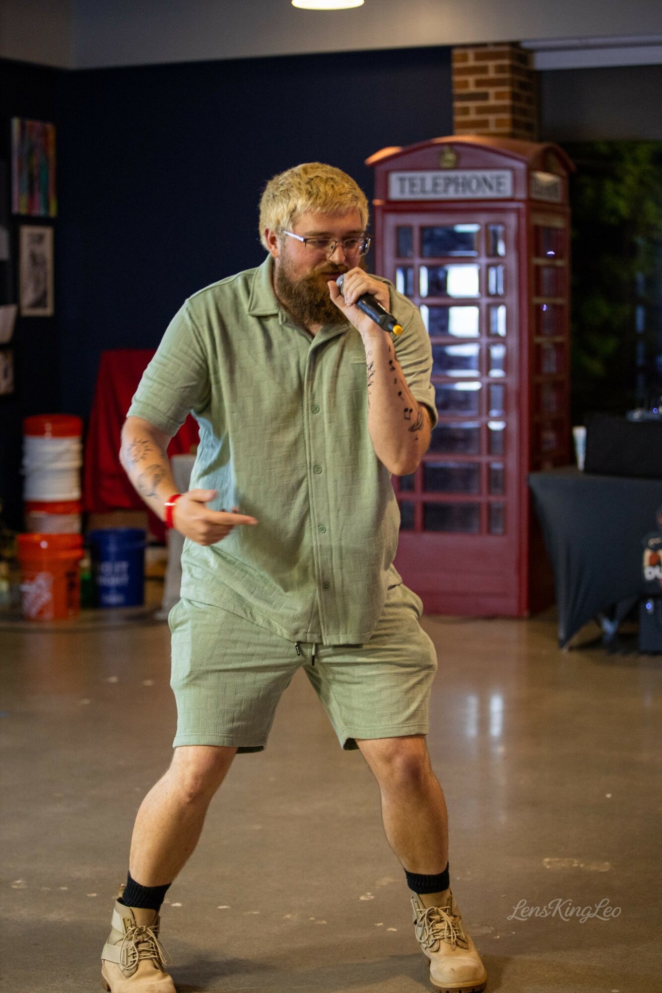 Person with glasses and beard holding microphone, standing indoors near a red telephone booth, wearing a green shirt and shorts.