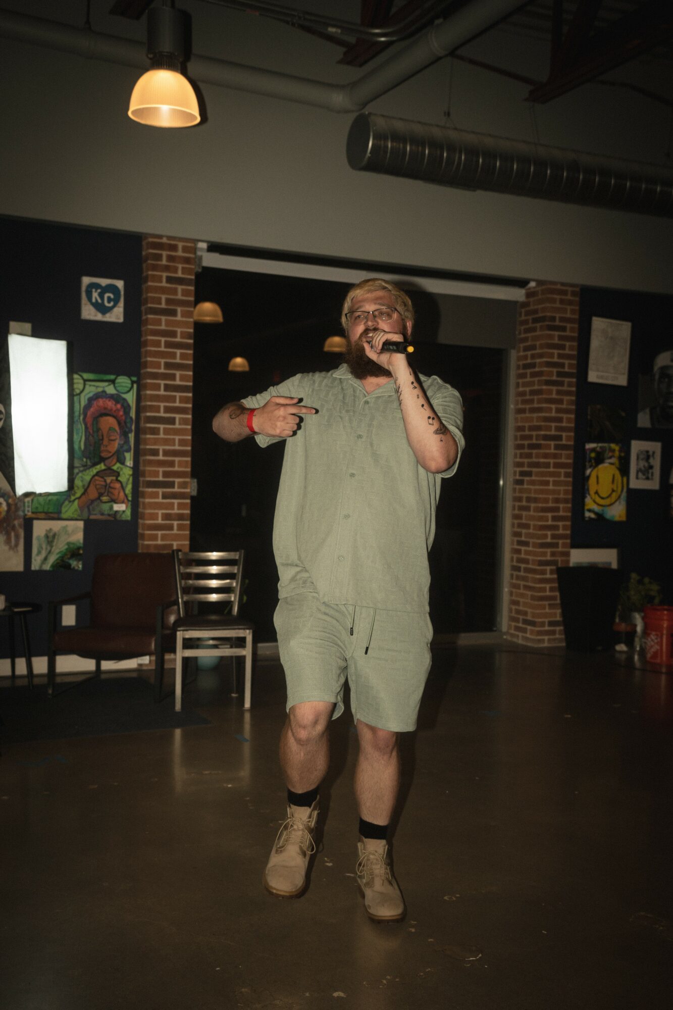 Person performing on stage with microphone, brick wall background, dim lighting, casual clothing, and sneakers.